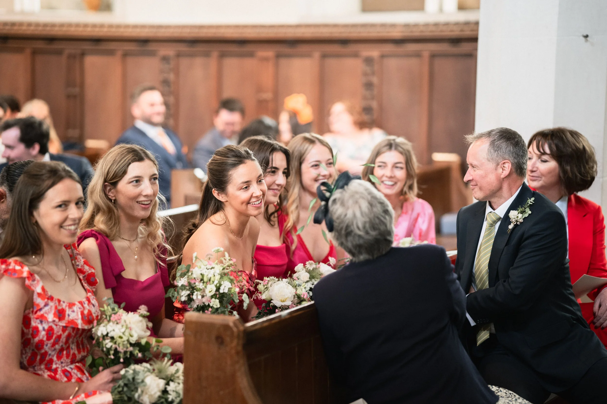 A wedding ceremony inside a church with smiling women seated in pews, listening to an older woman and a man in a suit speaking, near the bride dressed in pink and holding a bouquet of flowers.