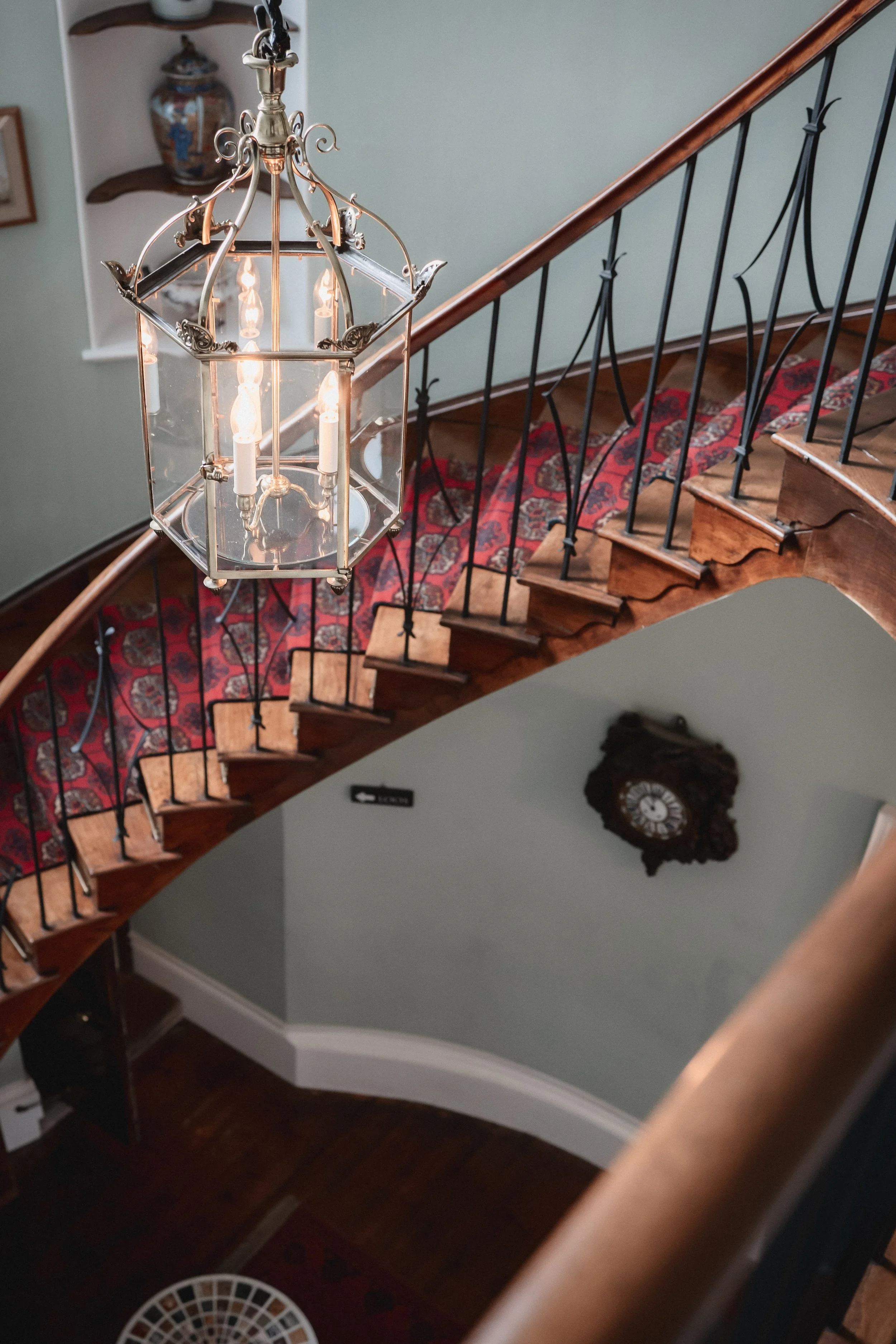 Upward view of a staircase with a decorative chandelier hanging above, a patterned rug on the stairs, and a clock on the wall below.