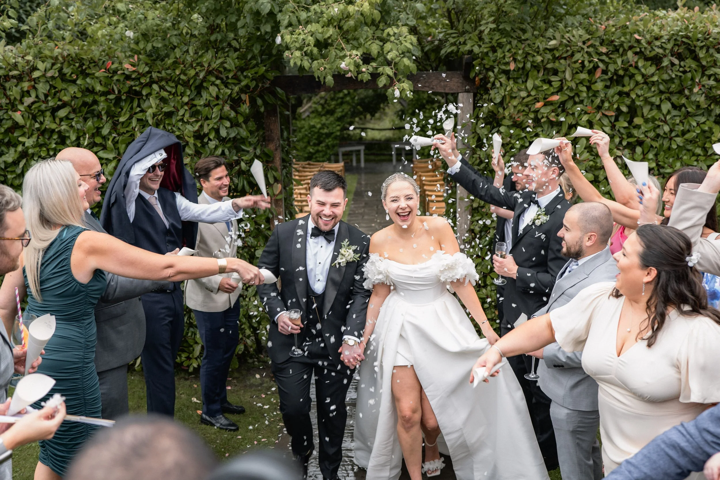 A bride and groom walking hand in hand through a crowd of wedding guests who are throwing confetti and celebrating outdoors.
