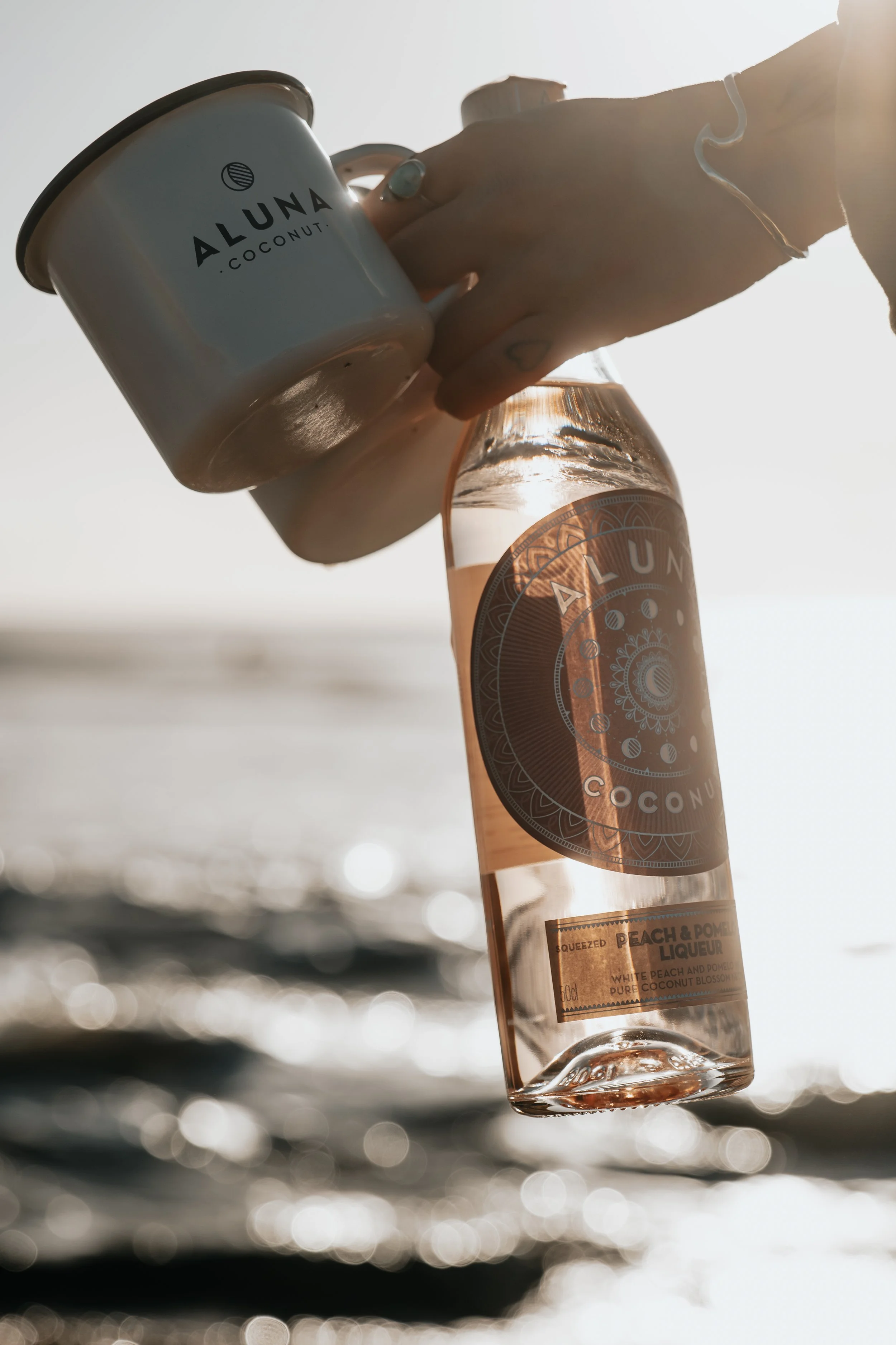 Person holding a jar of Aluna Coconut beverage and a bottle of Aluna Coconut Peach & Pomegranate Liqueur with a beach and water in the background during sunset.