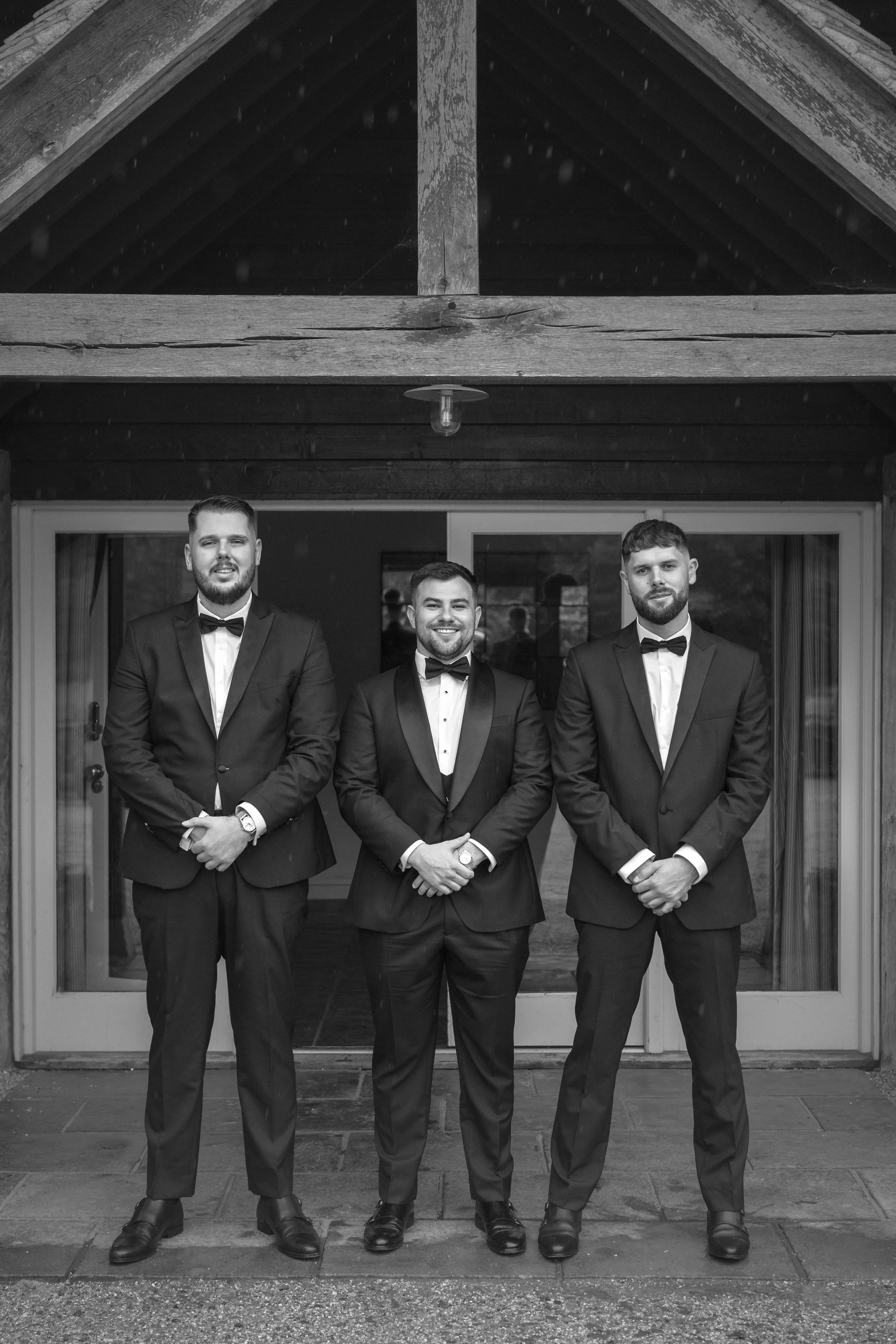 Three men in tuxedos standing in front of a house, smiling. It appears to be a formal event or wedding.