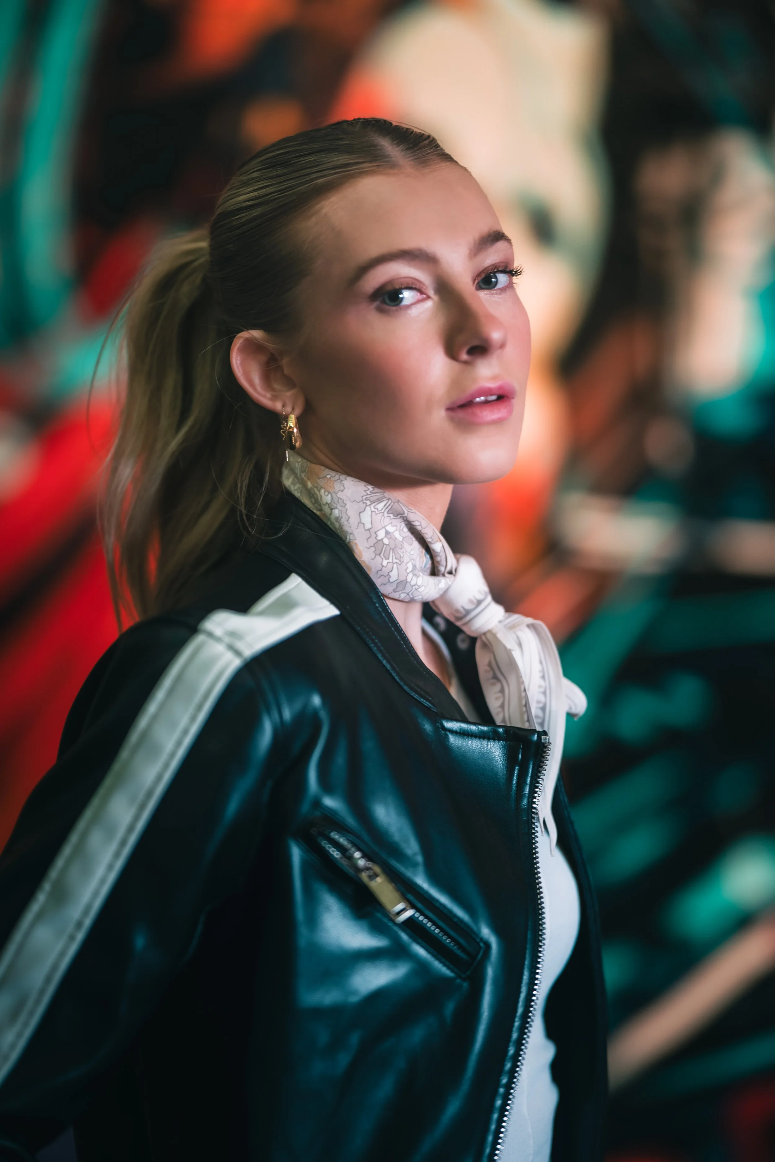 Portrait of a young woman with blonde hair pulled back, wearing a black leather jacket and a white scarf, looking at the camera with a blurred colorful background.