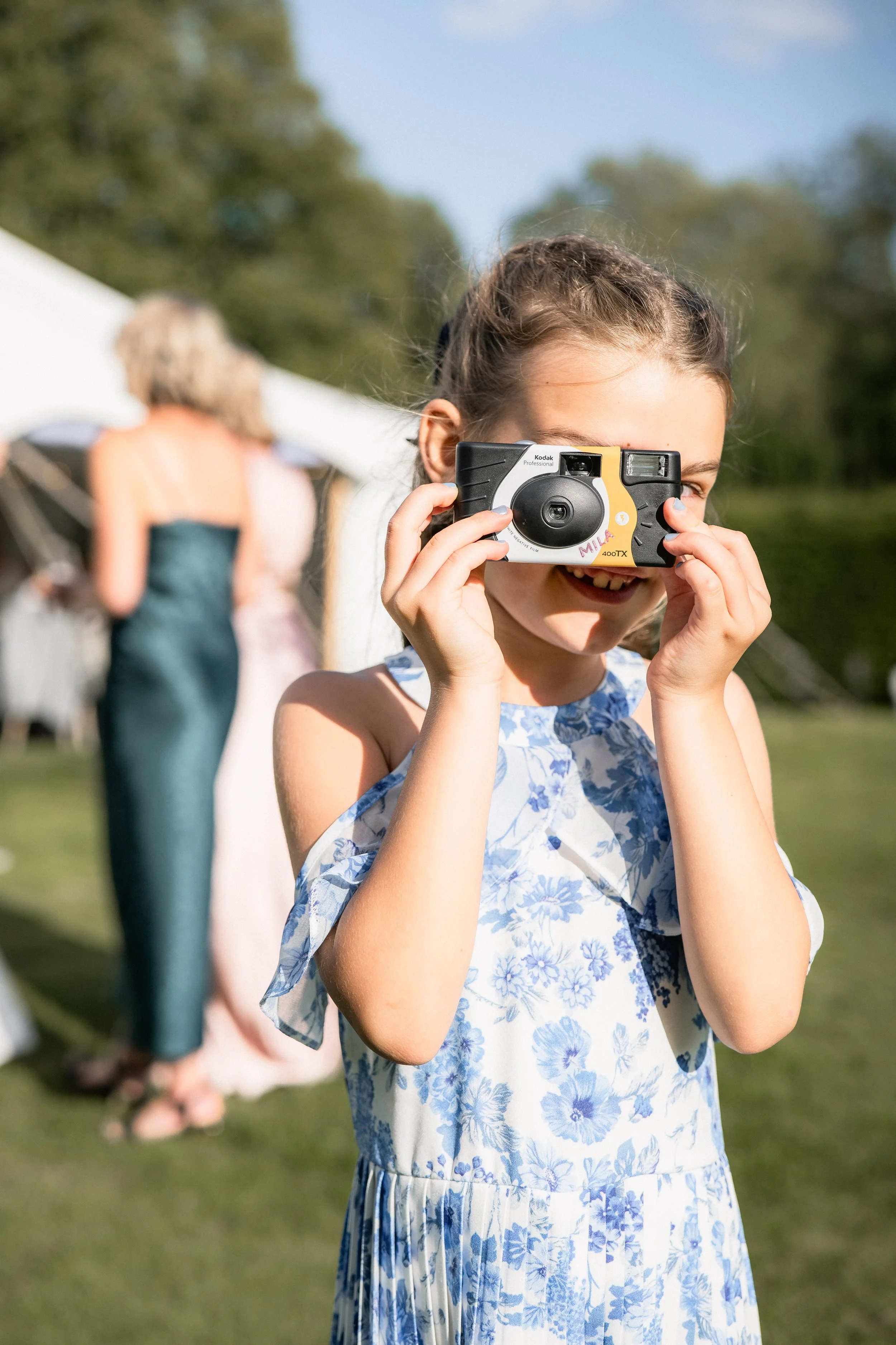 A young girl with braided hair wearing a floral dress, smiling, and holding a camera to her face outdoors during daytime.