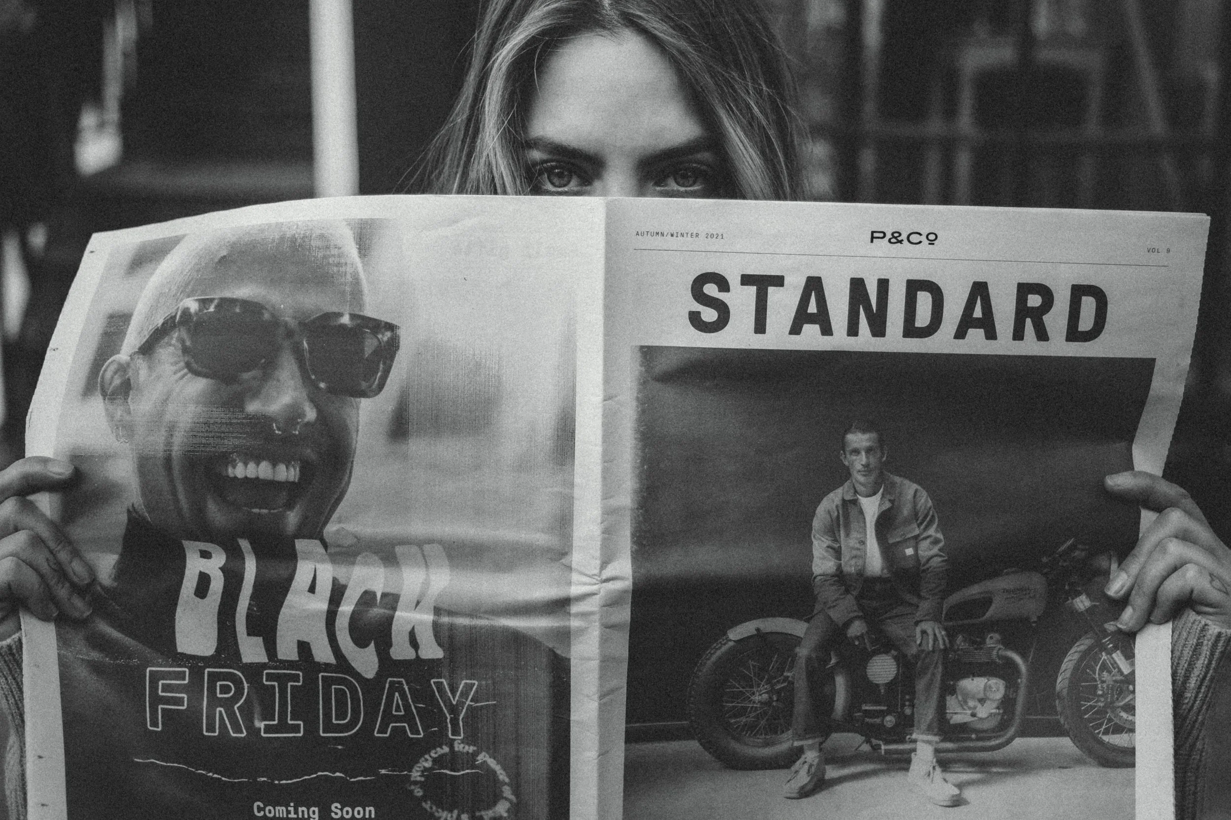 A woman with long hair holding a newspaper in front of her face, with only her eyes visible above the newspaper. The newspaper features a large image of a smiling man wearing sunglasses and a newsstand headline that reads 'STANDARD' and a photo of a 