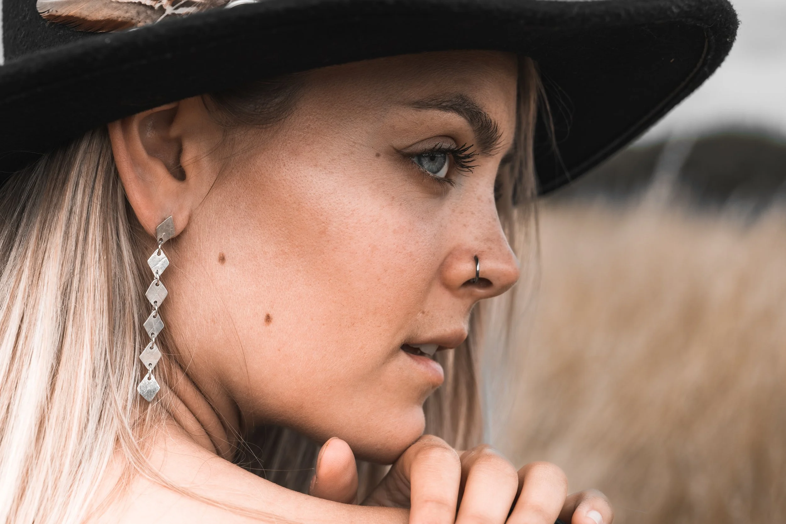 Close-up of a woman with blonde hair wearing a wide-brimmed black hat, silver drop earrings, a nose ring, and natural makeup, looking pensively into the distance outdoors.