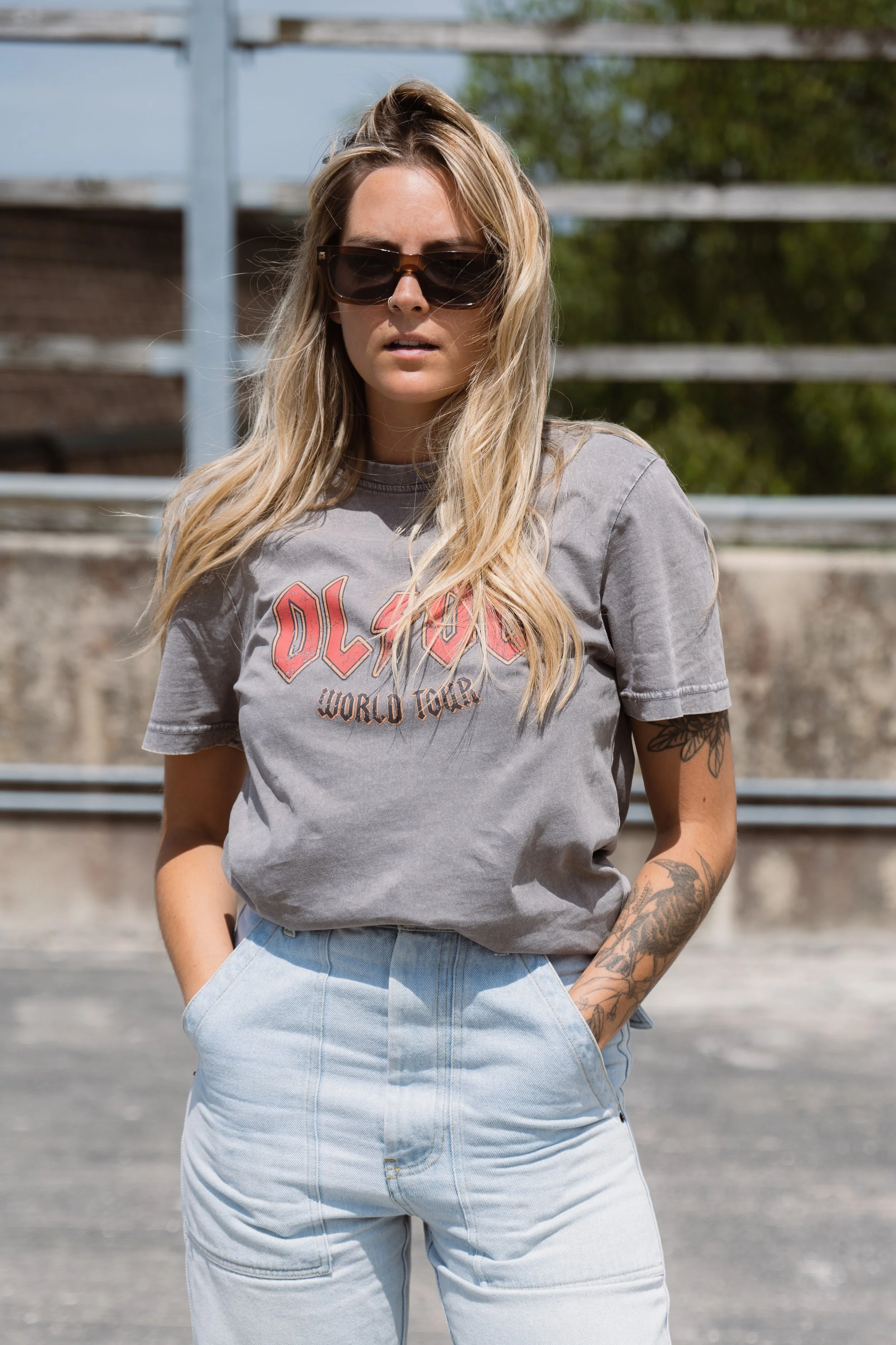 A young woman with blonde hair wearing sunglasses, a gray AC/DC T-shirt, and light blue jeans standing outdoors with her hands in her pockets.