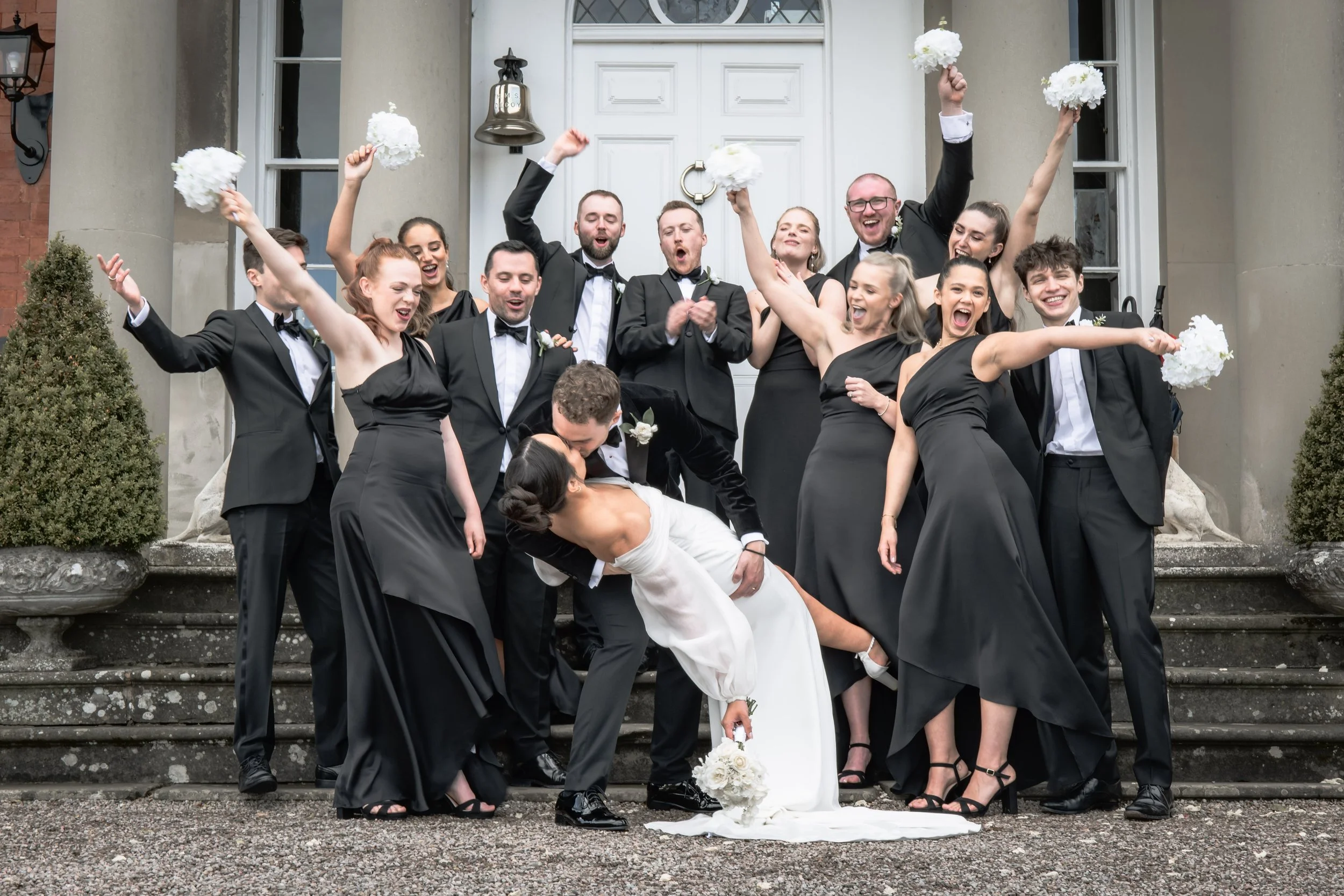 A group of wedding guests, dressed in formal black attire, celebrating on the steps outside a building. The bride, wearing a white dress, is being kissed by a groom who is bending down, and the guests are cheering, holding bouquets, and raising their