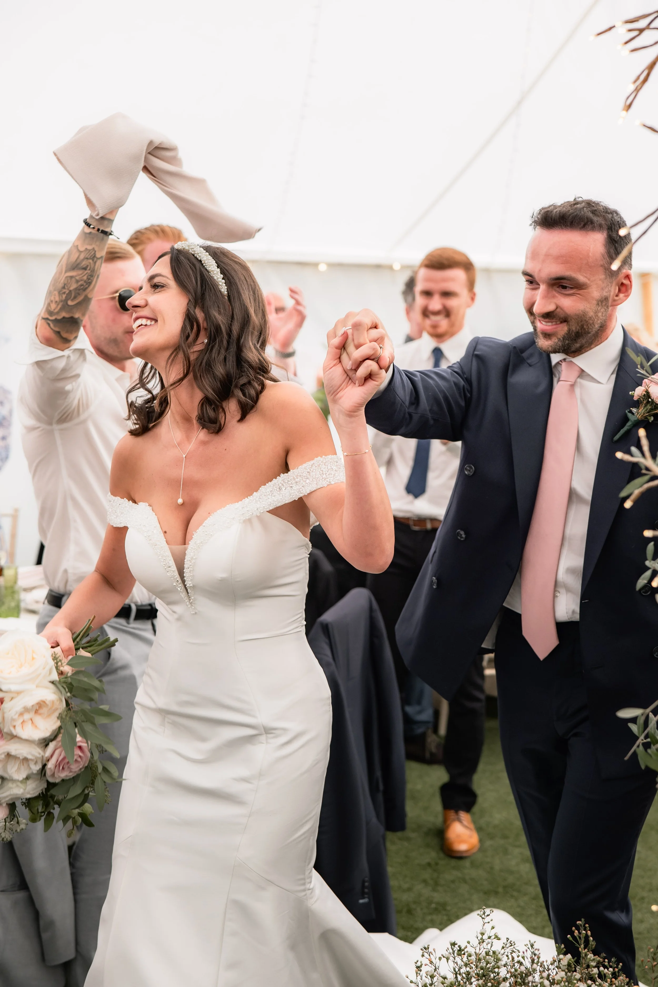 A bride and groom holding hands and smiling at a wedding reception, surrounded by celebrating guests.