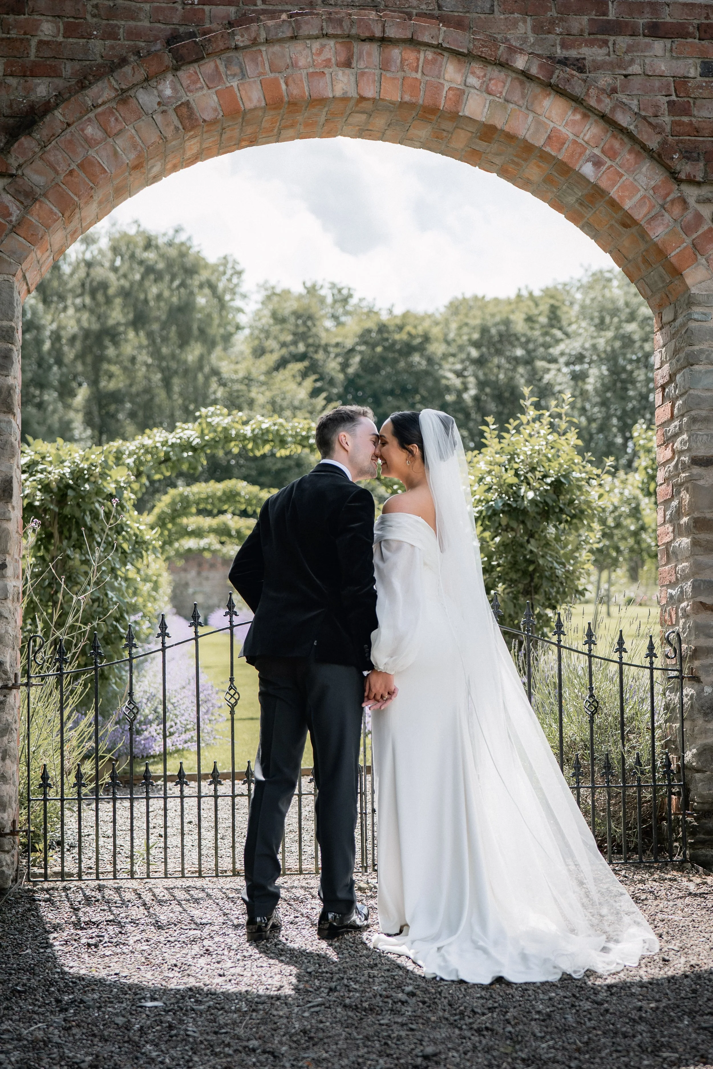 A bride and groom holding hands and touching foreheads under a brick archway, with a garden and trees in the background.