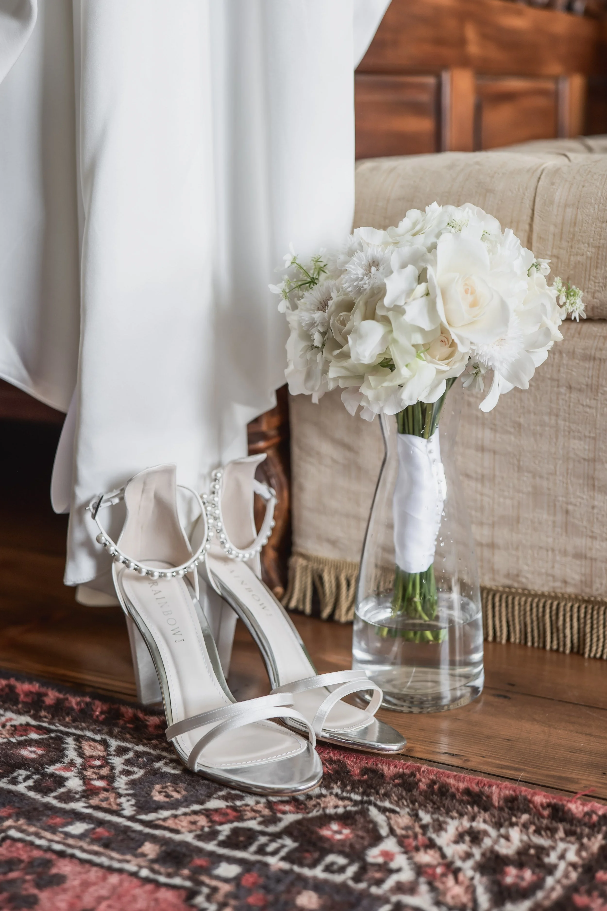 A pair of white high-heeled sandals with pearl ankle straps, a bouquet of white flowers in a glass vase, and a white garment hanging nearby, all on a wooden floor with a decorative rug.