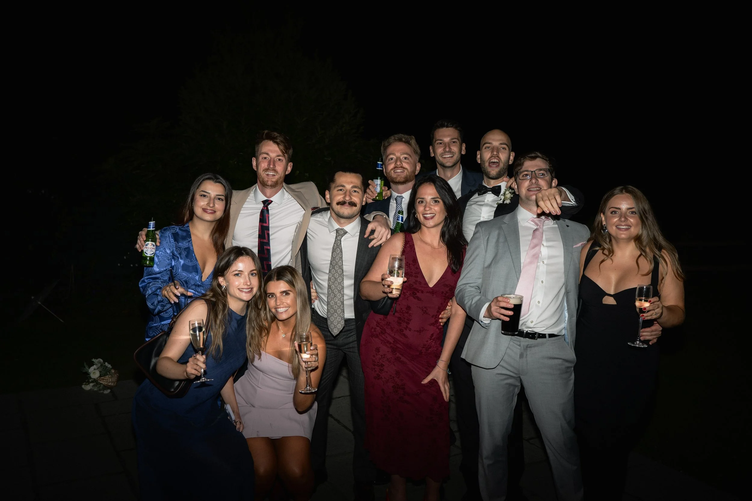 Group of people at a party or celebration, dressed in formal attire, holding drinks, smiling, at night outdoors.