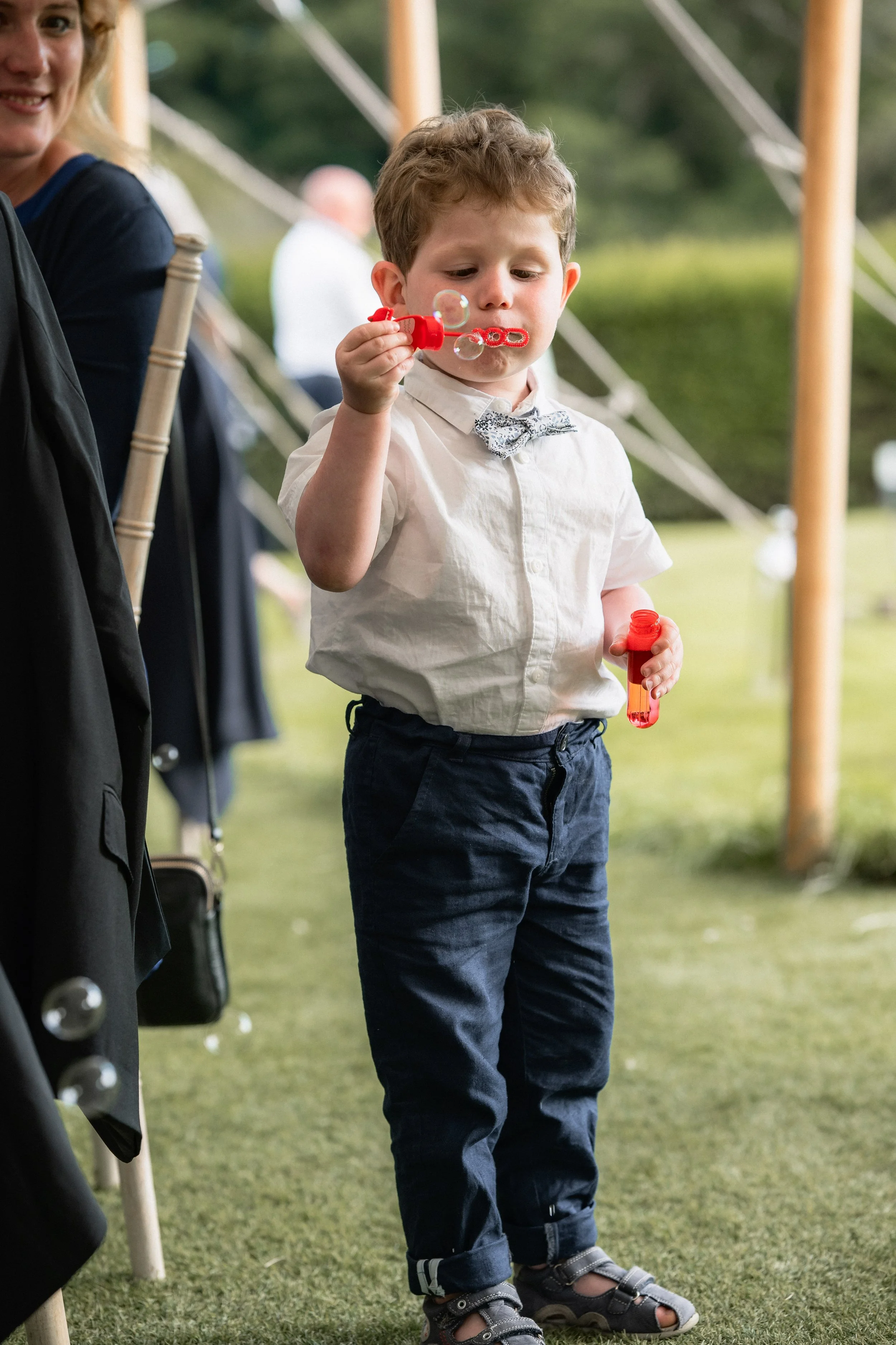 A young boy in a white shirt and dark pants blows bubbles at an outdoor event.