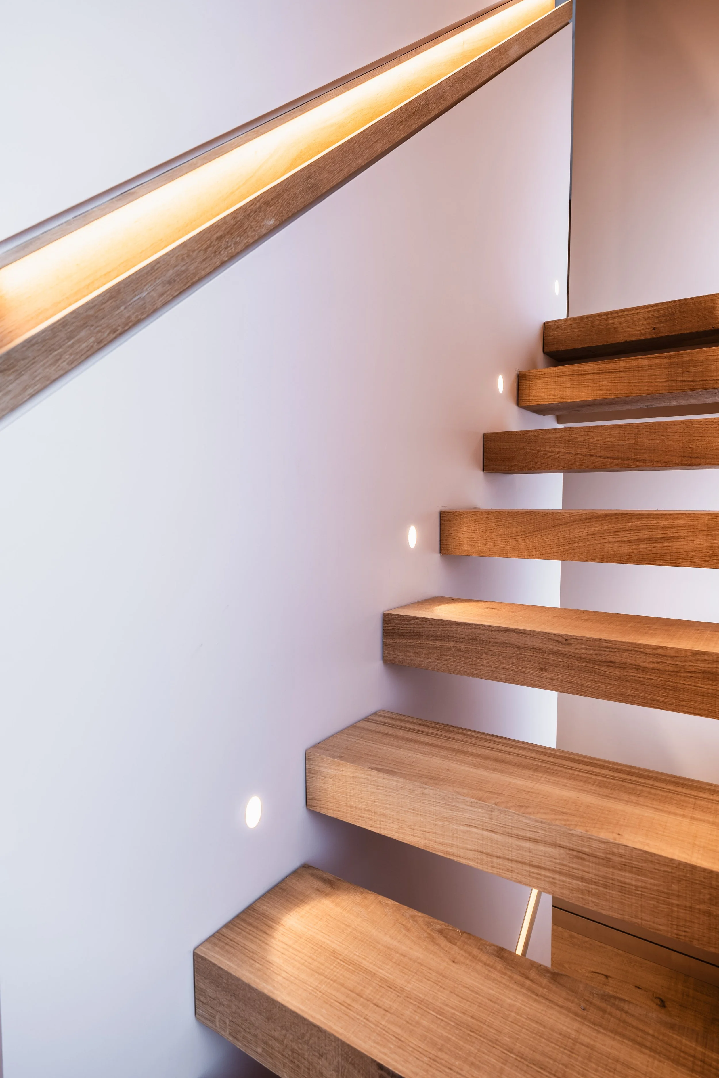 Close-up of a modern wooden staircase with embedded lighting on the white wall beside it.