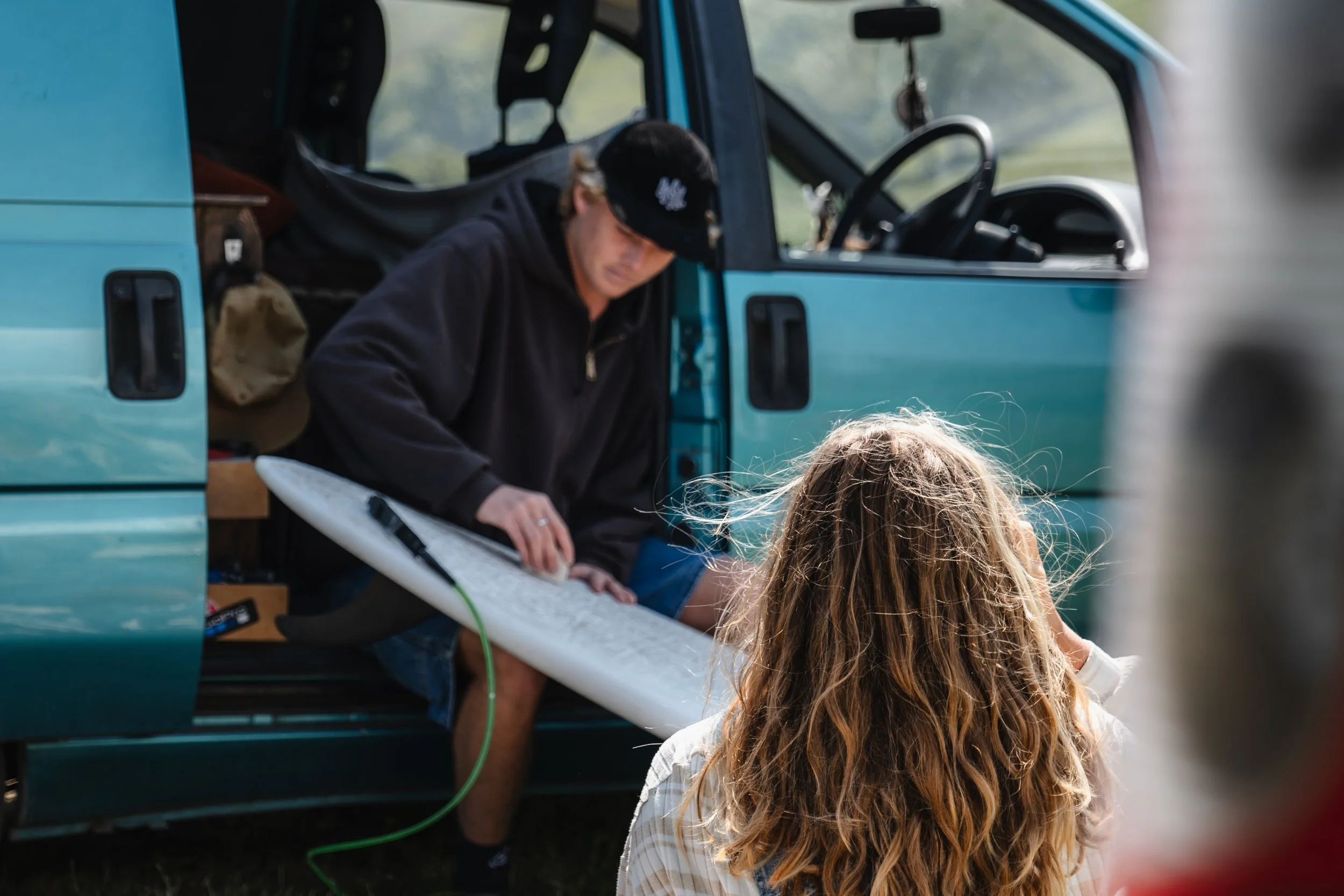 A man is sitting in the open doorway of a teal van, signing a surfboard, while a woman with long curly hair watches nearby.