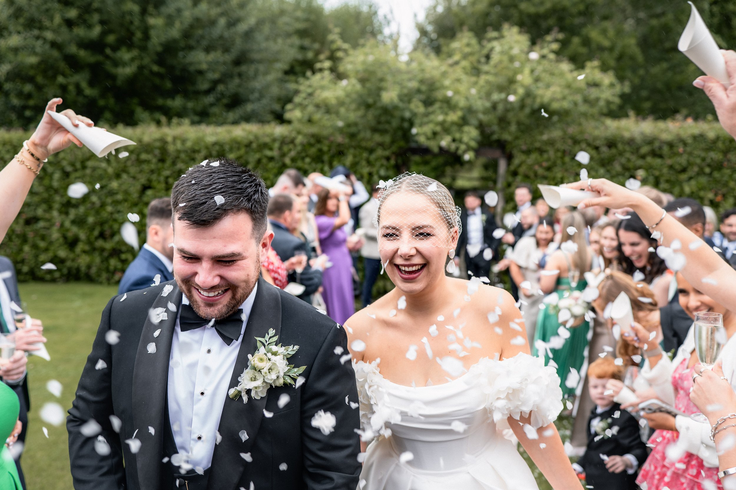 Bride and groom smiling as guests throw confetti during wedding celebration outdoors.
