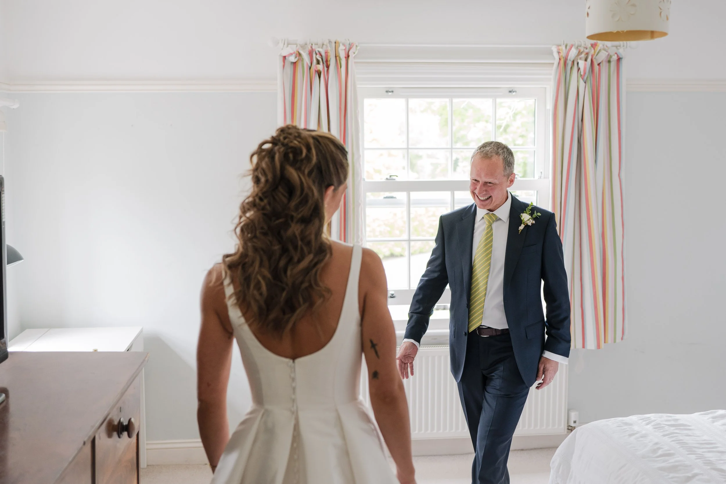 A bride facing an older man, possibly her father, in a bright room with a window and striped curtains, both smiling.