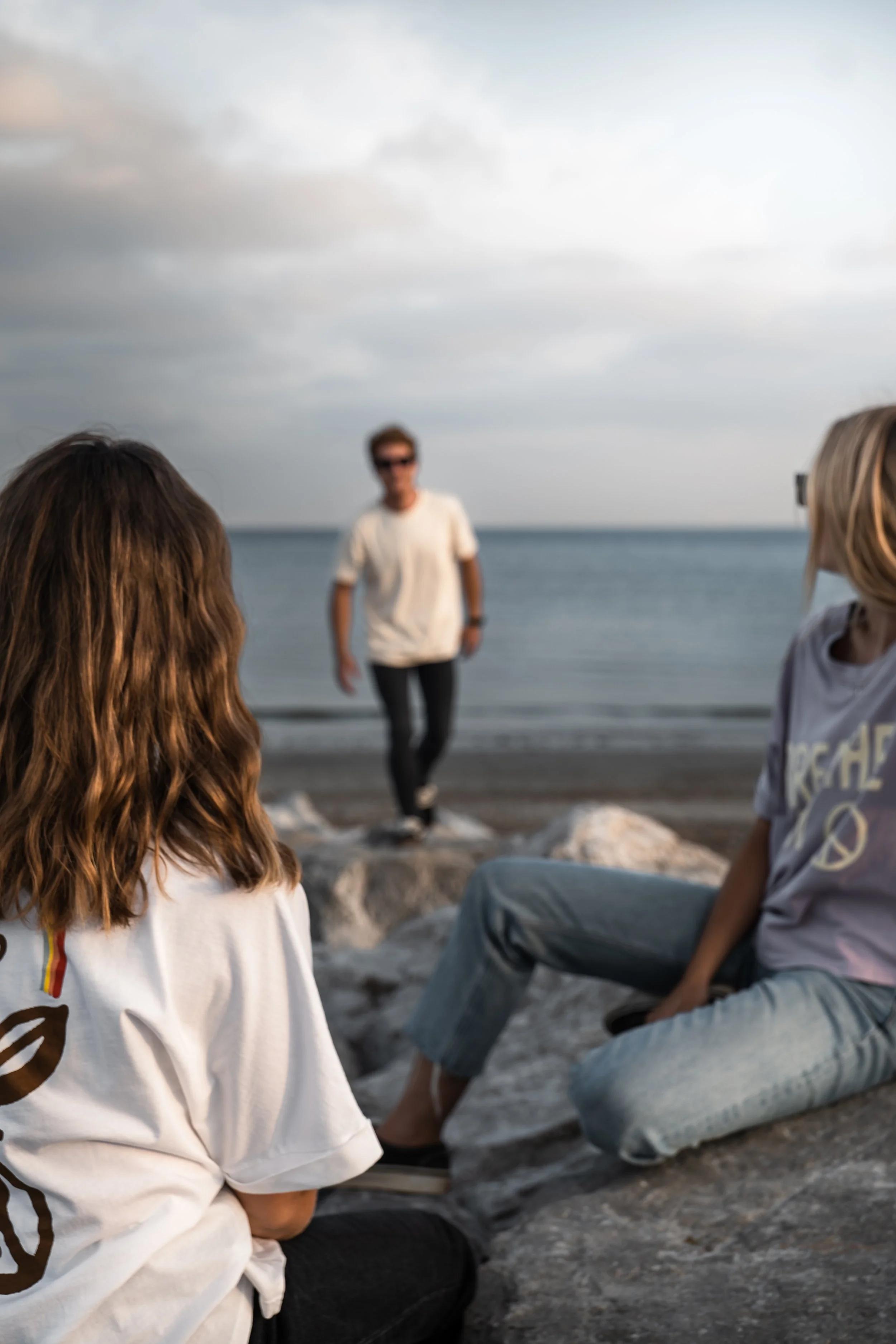 A woman and a girl sitting on rocks at the beach watching a woman walking towards them by the water during cloudy weather.