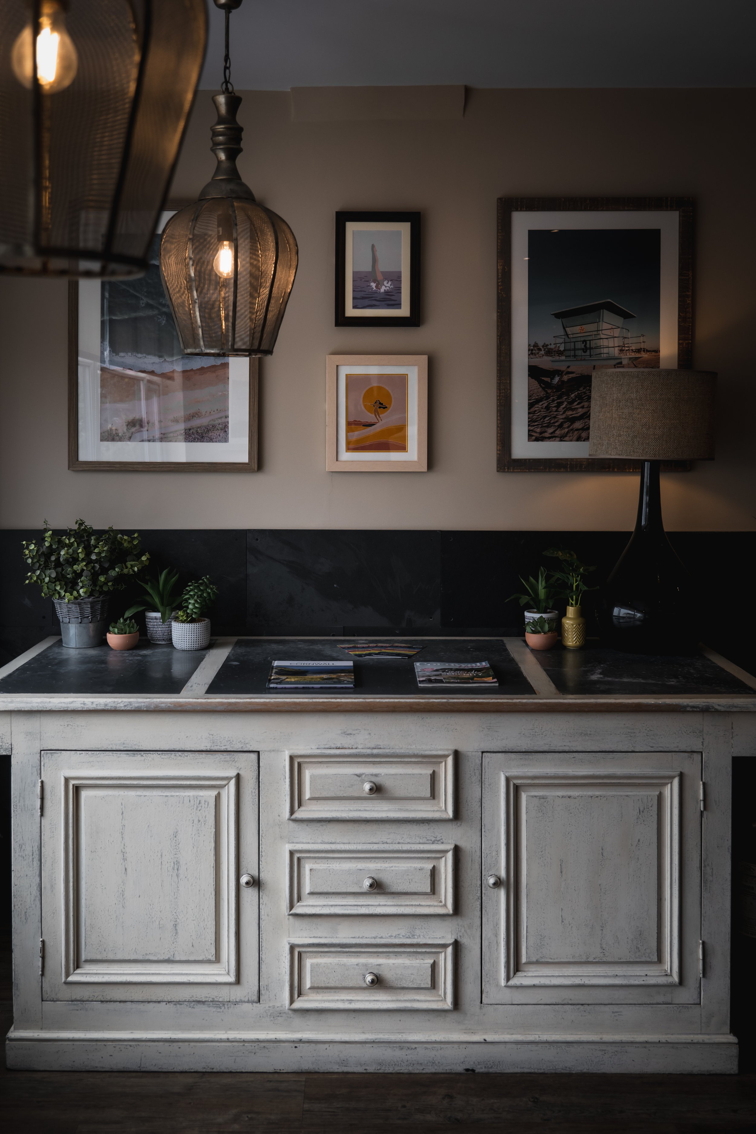 Vintage white wooden sideboard with three drawers and cabinet doors, topped with potted plants and magazines, in a dimly lit room with framed pictures on the wall and hanging lamps.