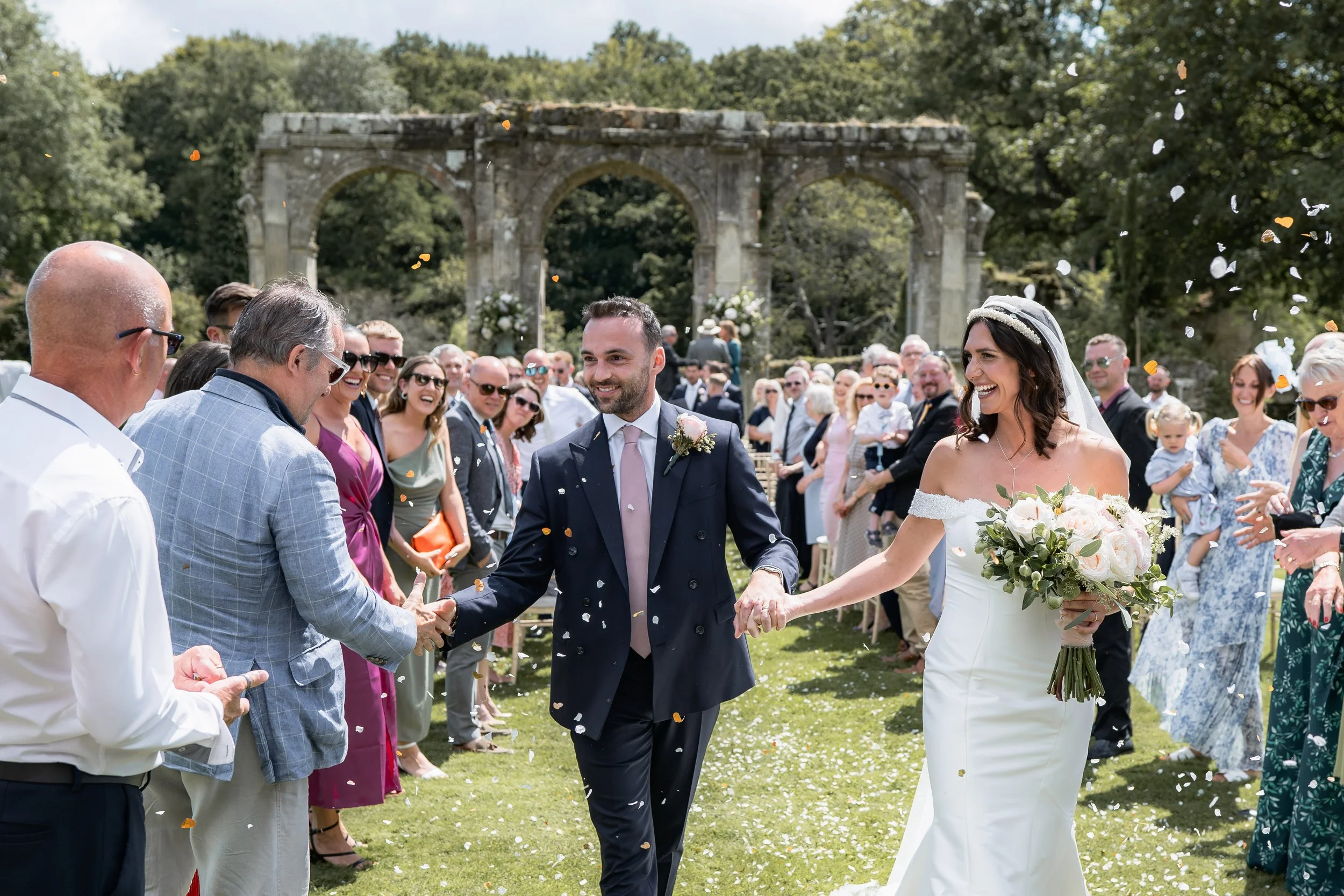 Bride and groom smiling and holding hands as they walk through a shower of confetti at an outdoor wedding ceremony, surrounded by guests in formal attire, with a stone arch and lush greenery in the background.