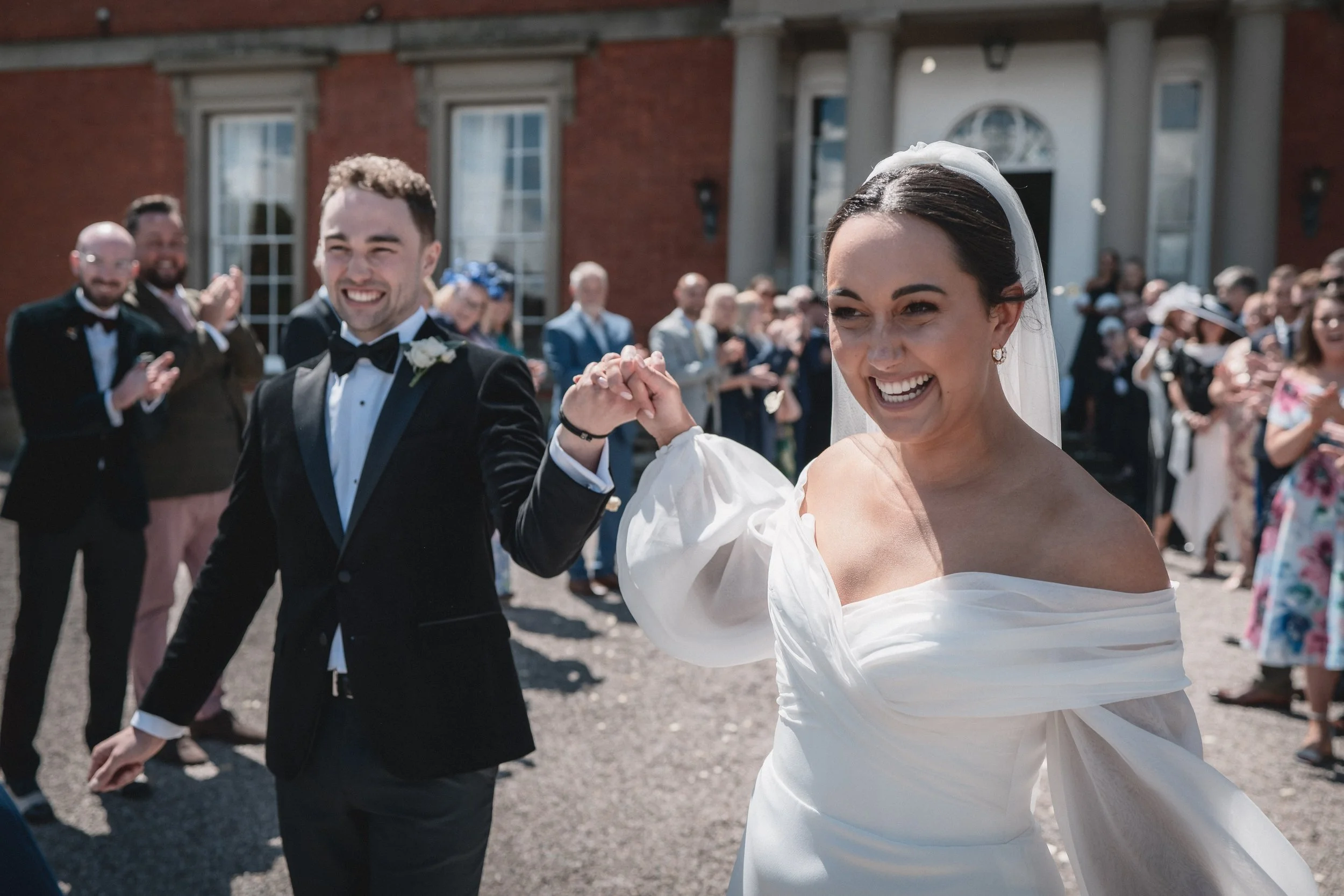 A bride and groom smiling and holding hands at their wedding outdoors, surrounded by guests.