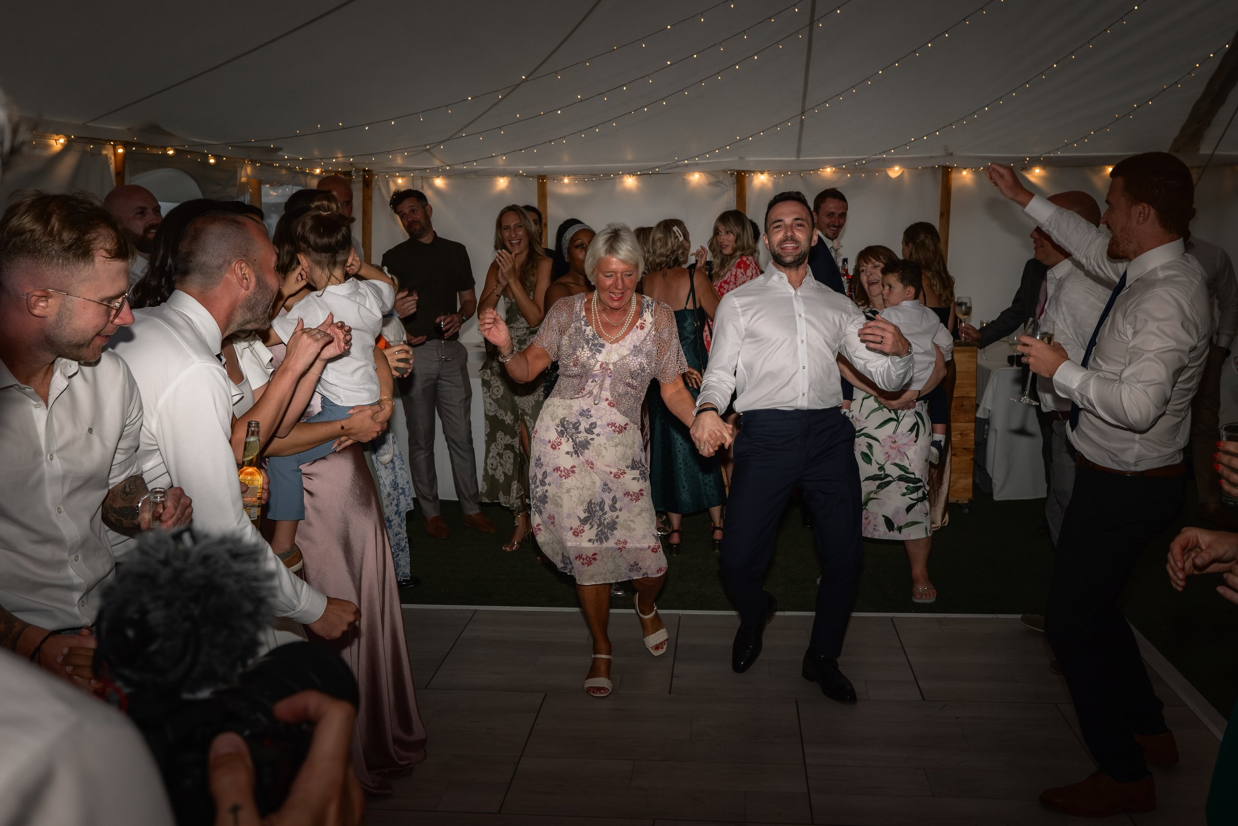 People dancing and celebrating at a wedding reception under a tent decorated with string lights.