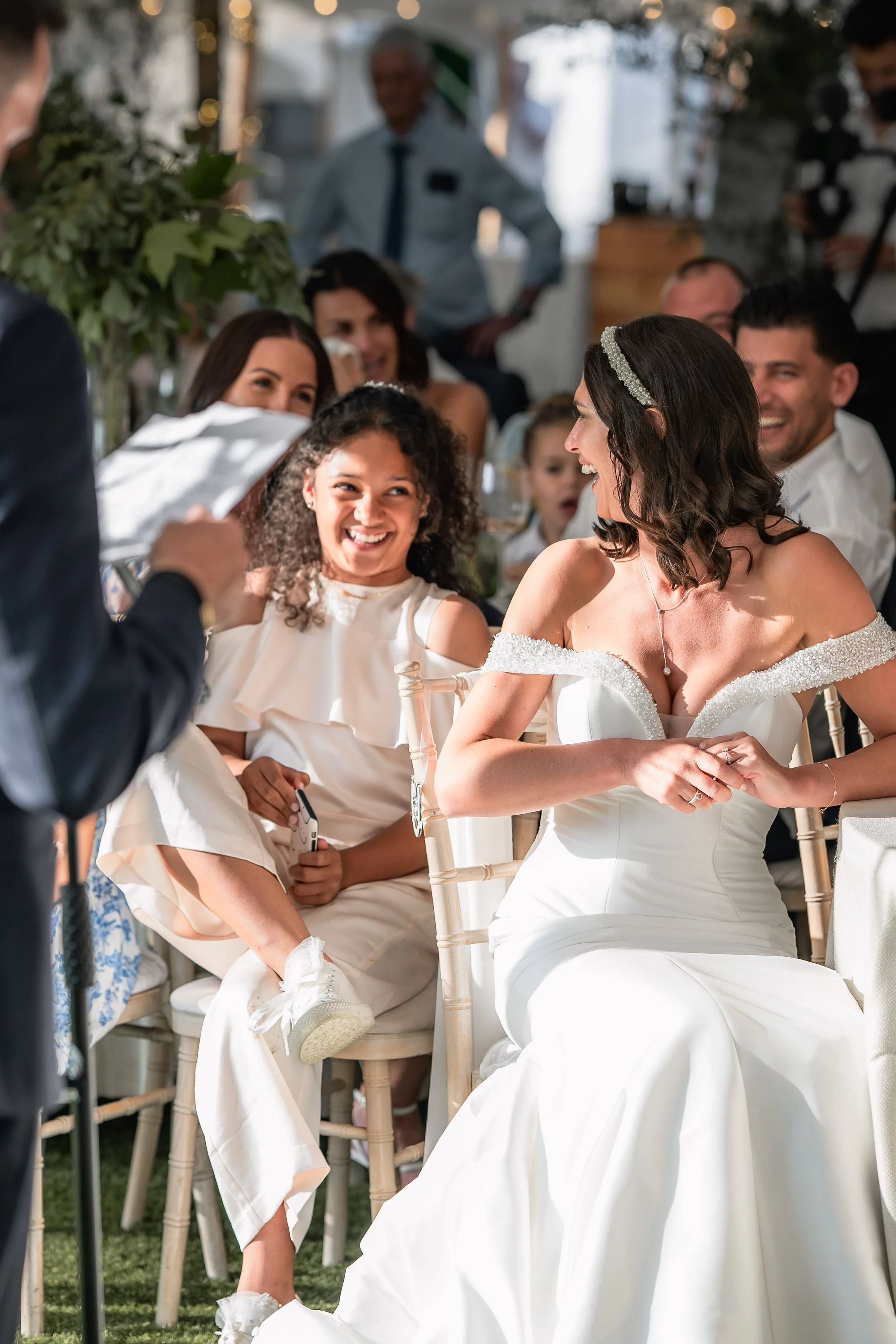 A bride in a white wedding dress laughing and holding hands with a woman sitting next to her at a wedding reception. Guests are smiling and enjoying the moment.