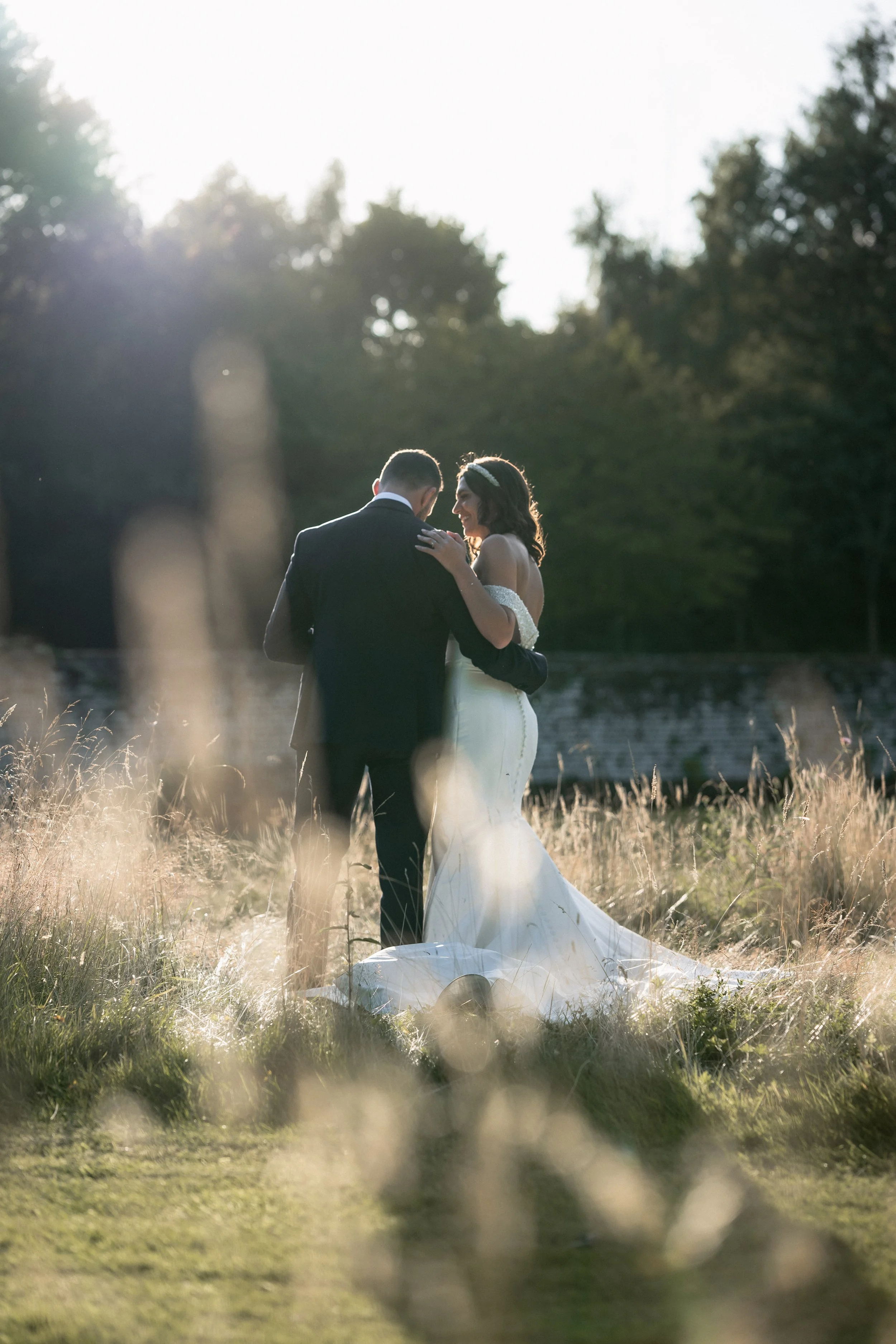 A bride and groom standing together outdoors in a grassy field, sharing a moment at sunset with trees in the background.