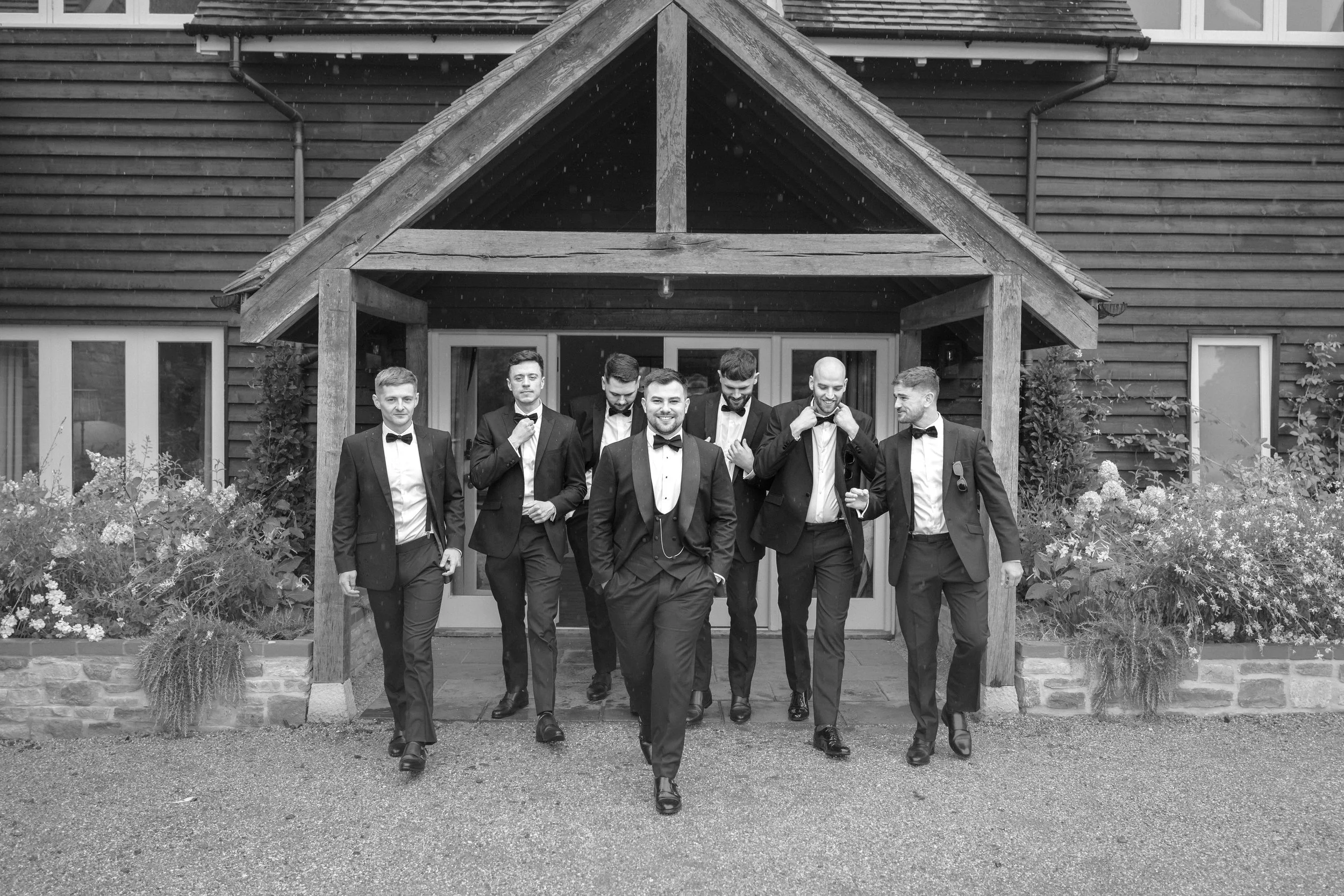A group of eight men in suits and bow ties walking out of a building with a wooden facade, some adjusting their bow ties, smiling and looking confident.