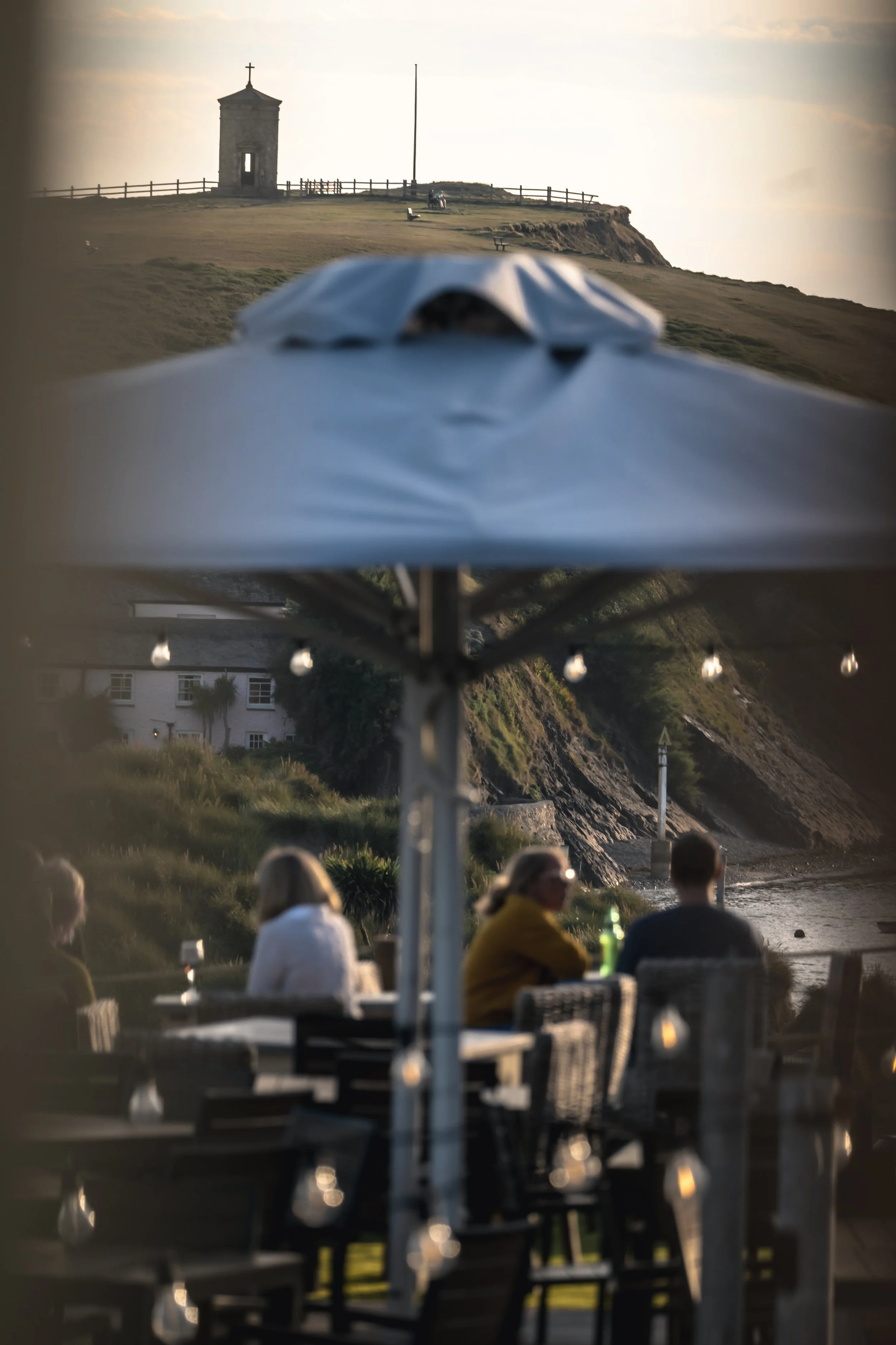 People sitting at an outdoor restaurant near the coast with a view of a grassy hill, lighthouse, and a building in the background, during sunset.