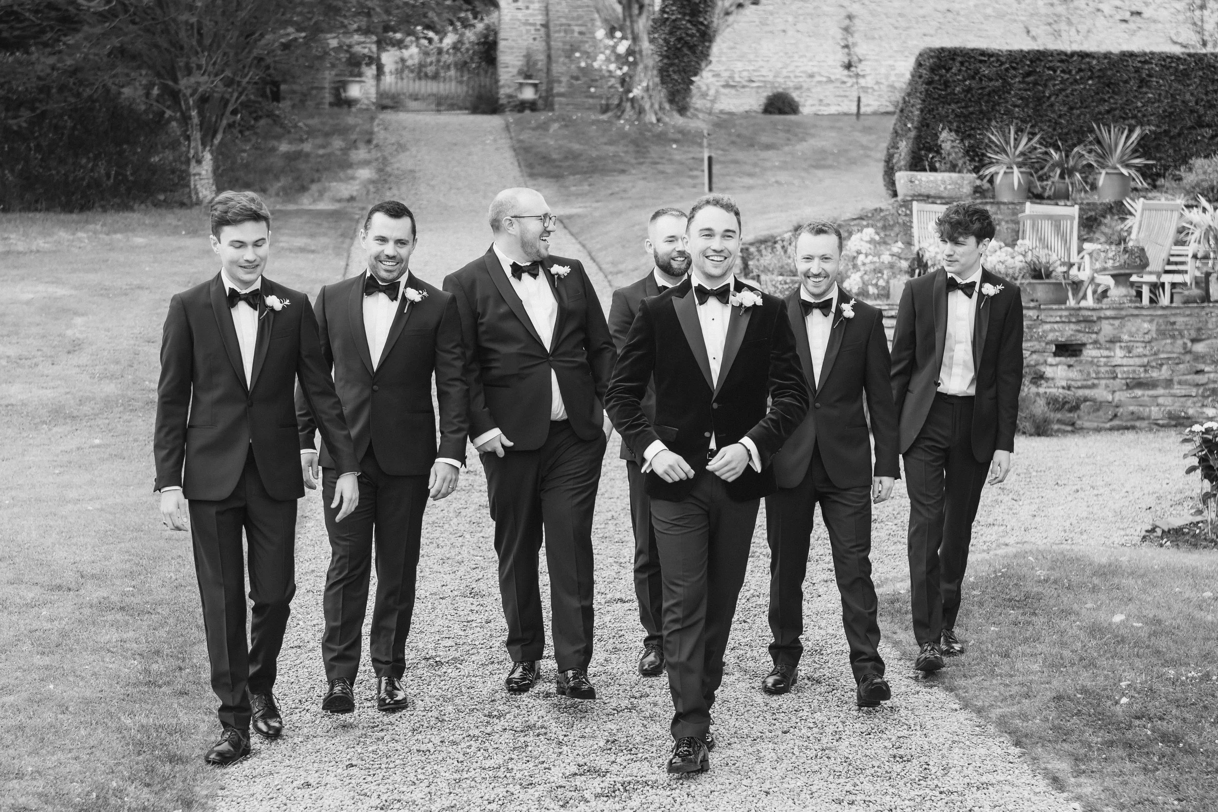 A group of seven men in tuxedos walking outdoors in a garden, smiling and laughing.
