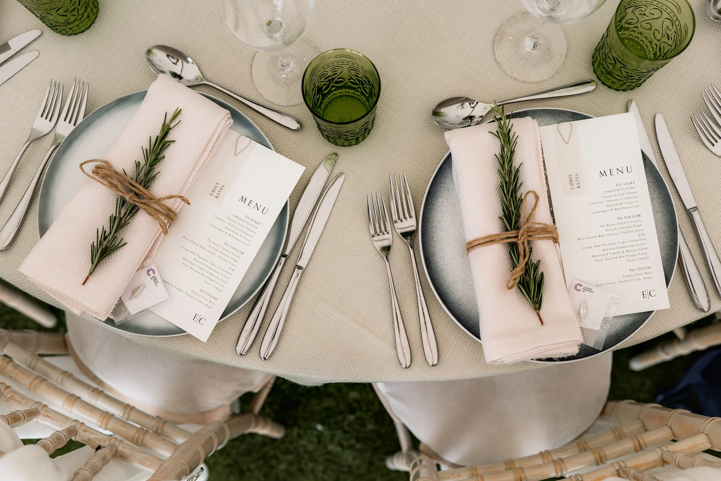Elegant table setting with two place settings, each featuring a black plate, a pink napkin topped with a sprig of rosemary tied with twine, menus, and silverware, along with green glasses and stemware on a cream-colored tablecloth.