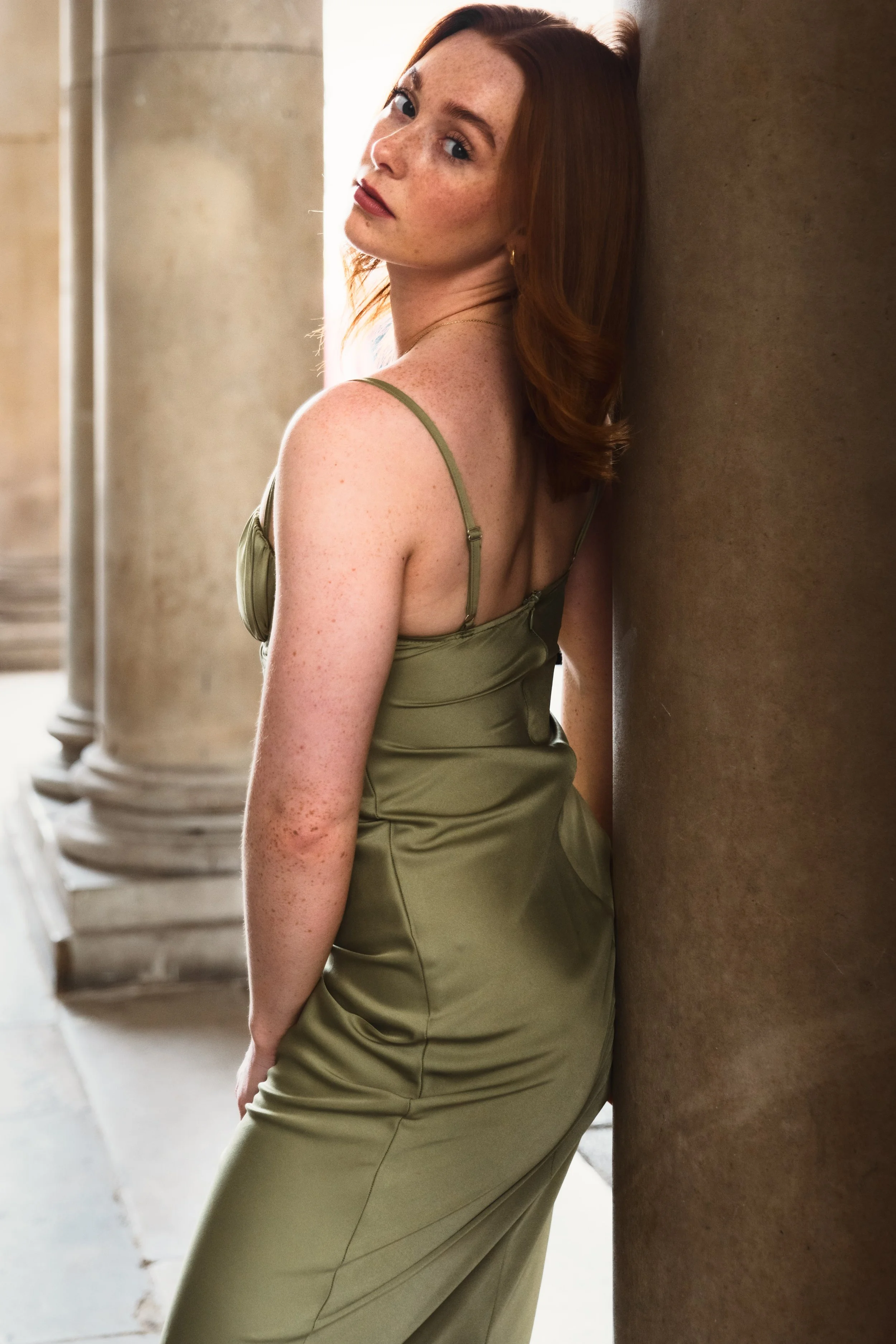 A woman with red hair and freckles leaning against a large stone column, wearing a satin olive green dress with spaghetti straps, looking toward the camera.