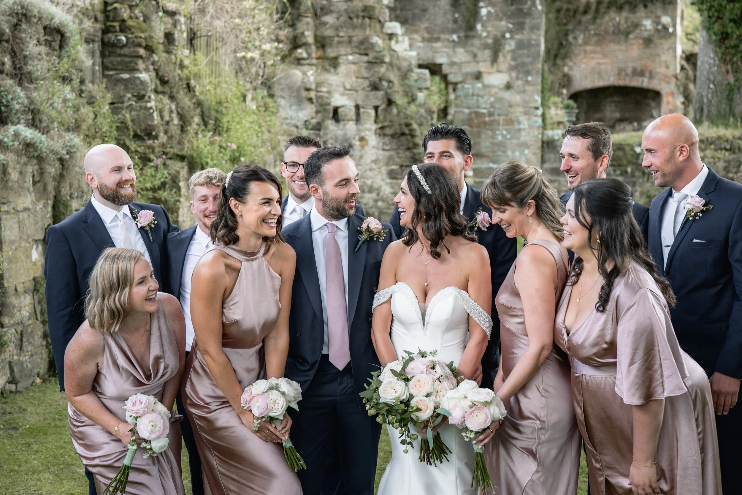 A wedding party outdoors with four women in mauve dresses and six men in dark suits, gathered around a bride in a white wedding gown holding a bouquet, and a groom in a dark suit with a pink tie, all smiling and looking at each other.