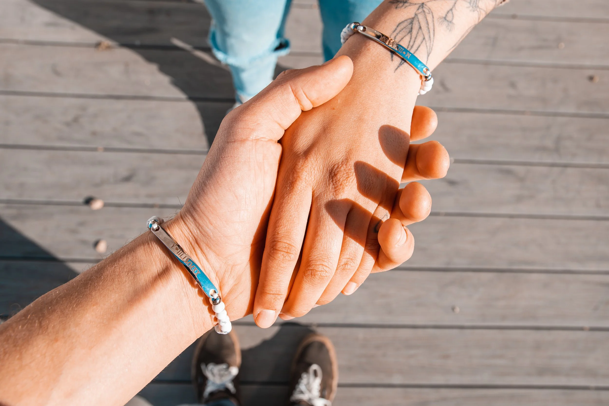 Two people are holding hands, with the person on the left wearing a bracelet and the person on the right wearing a bracelet and a ring. The background shows wooden planks and the photo is taken from above, with shoes visible at the bottom.
