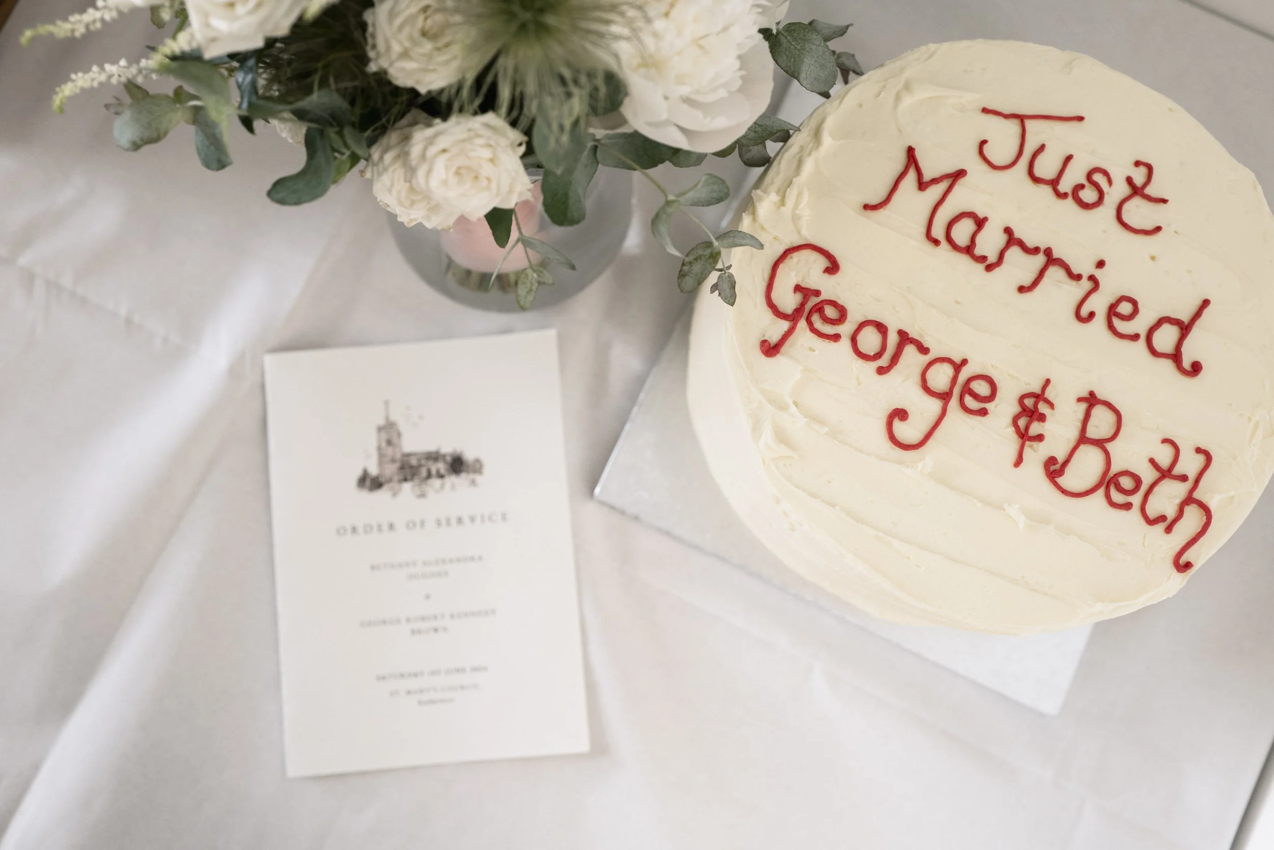 A white birthday cake with red piped writing that says 'Just Married George & Beth,' a floral arrangement in a vase, and a printed order of service on a white tablecloth.