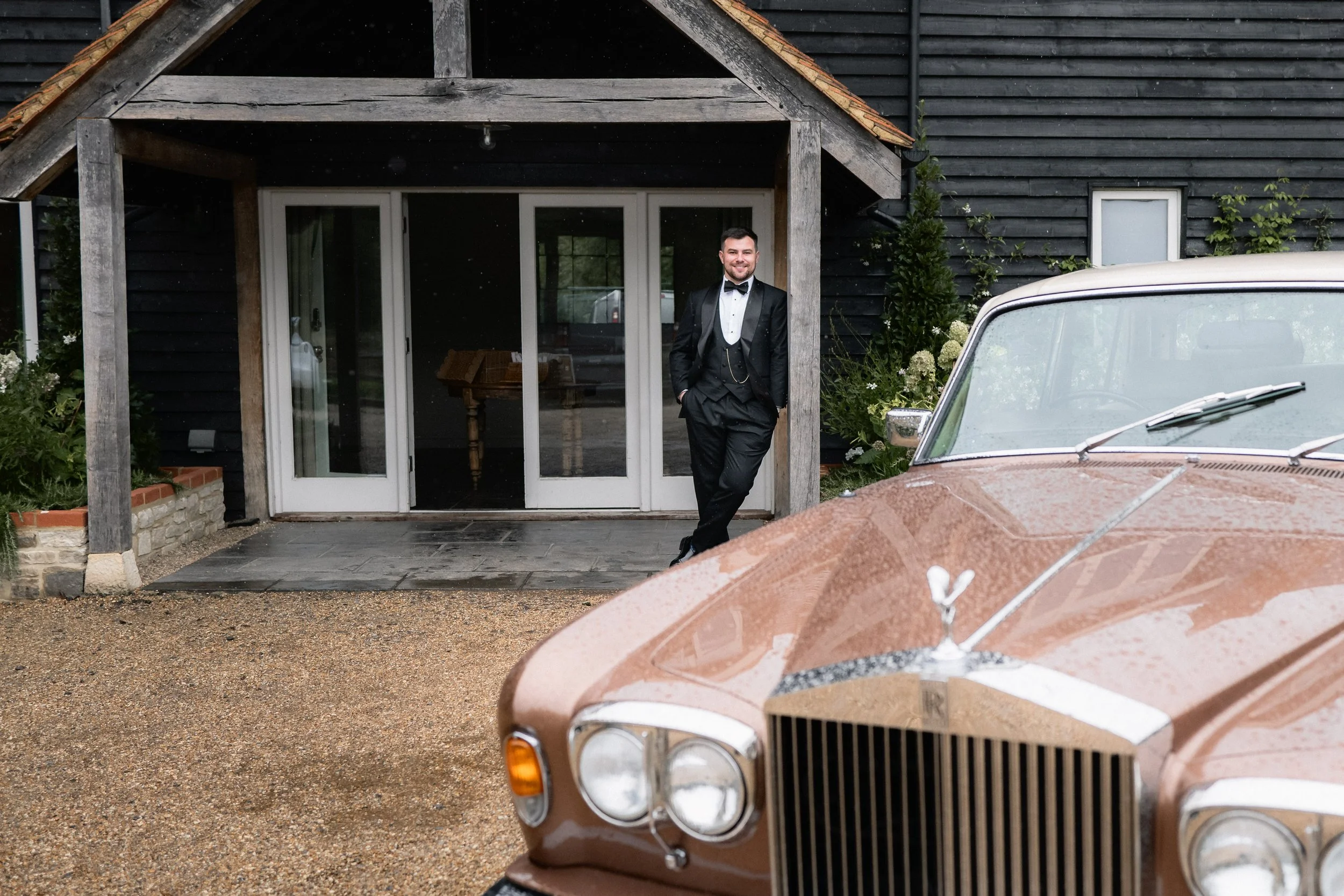 A man in a tuxedo standing by a vintage brown Rolls Royce car in front of a black wooden house with a porch.