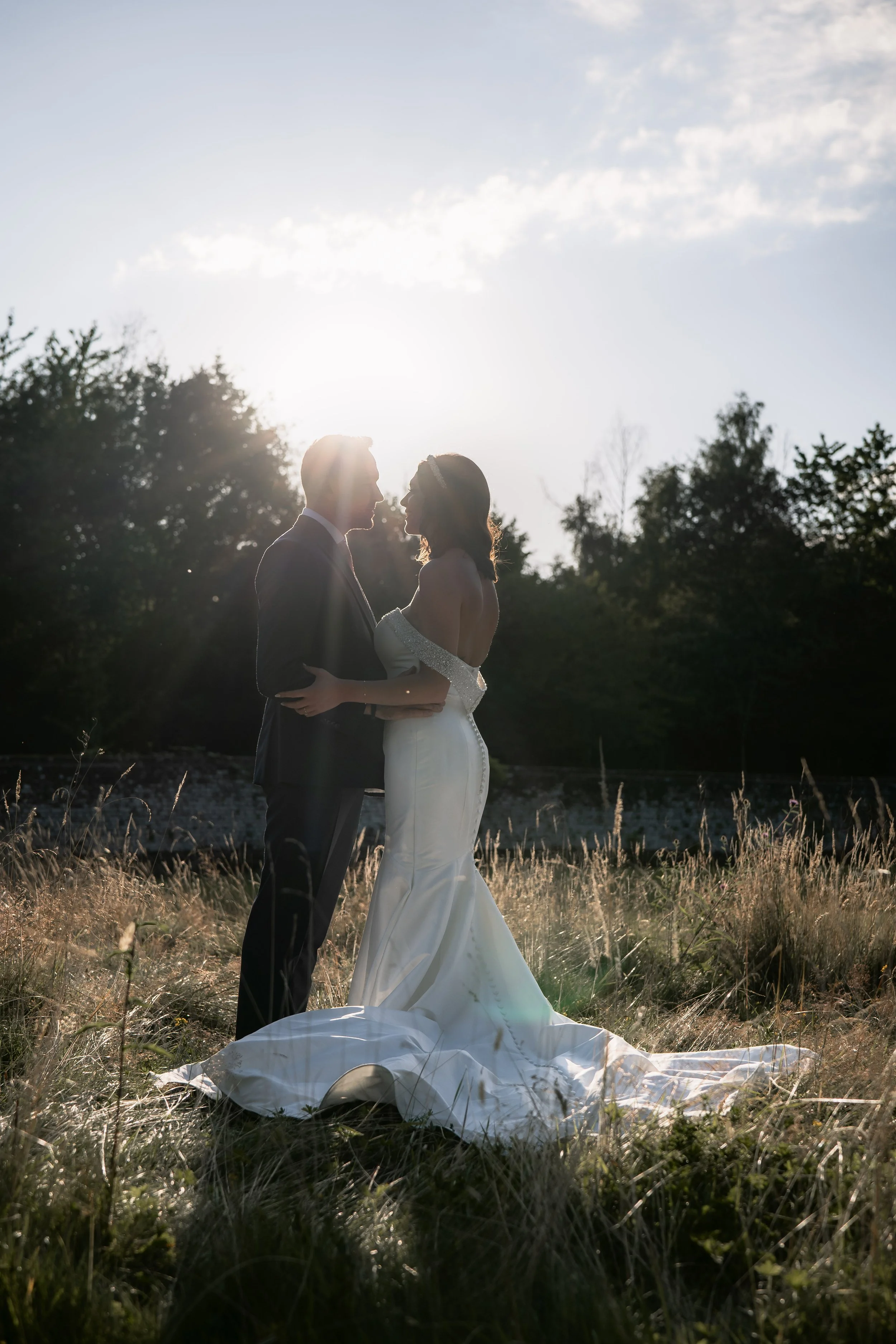 A bride and groom standing closely together outdoors during sunset, silhouetted against the sky, sharing a moment in their wedding attire.