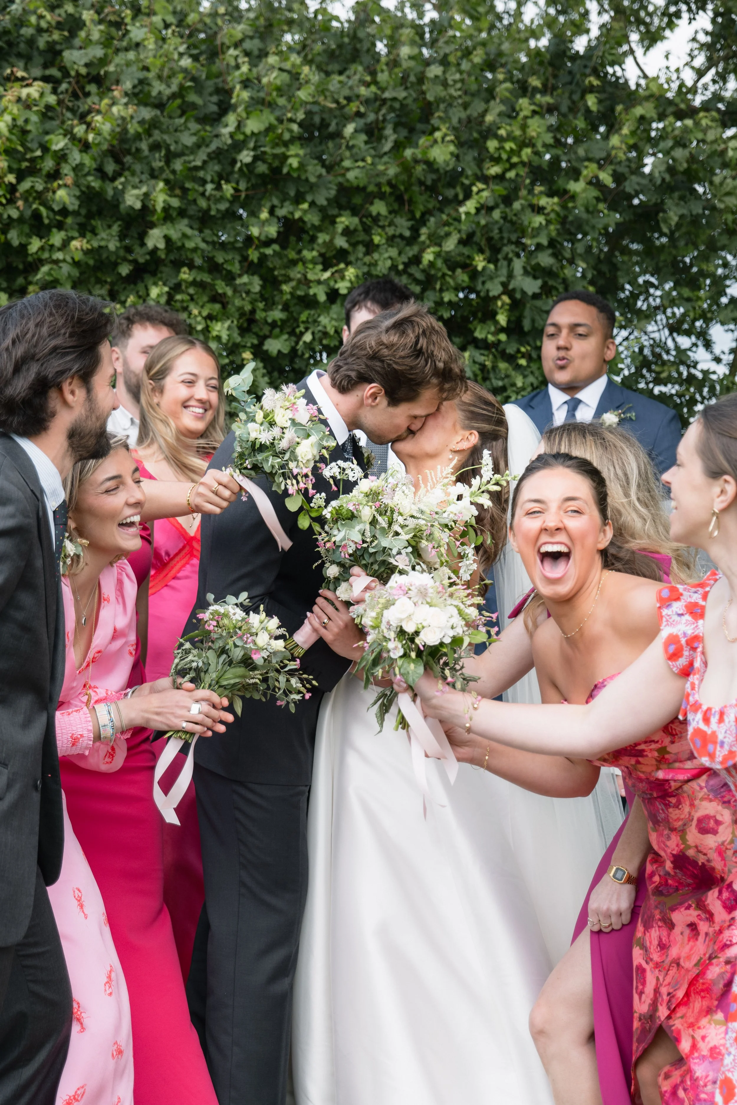 A group of people celebrating a wedding outdoors, with a couple kissing in the center surrounded by friends holding bouquets and smiling.