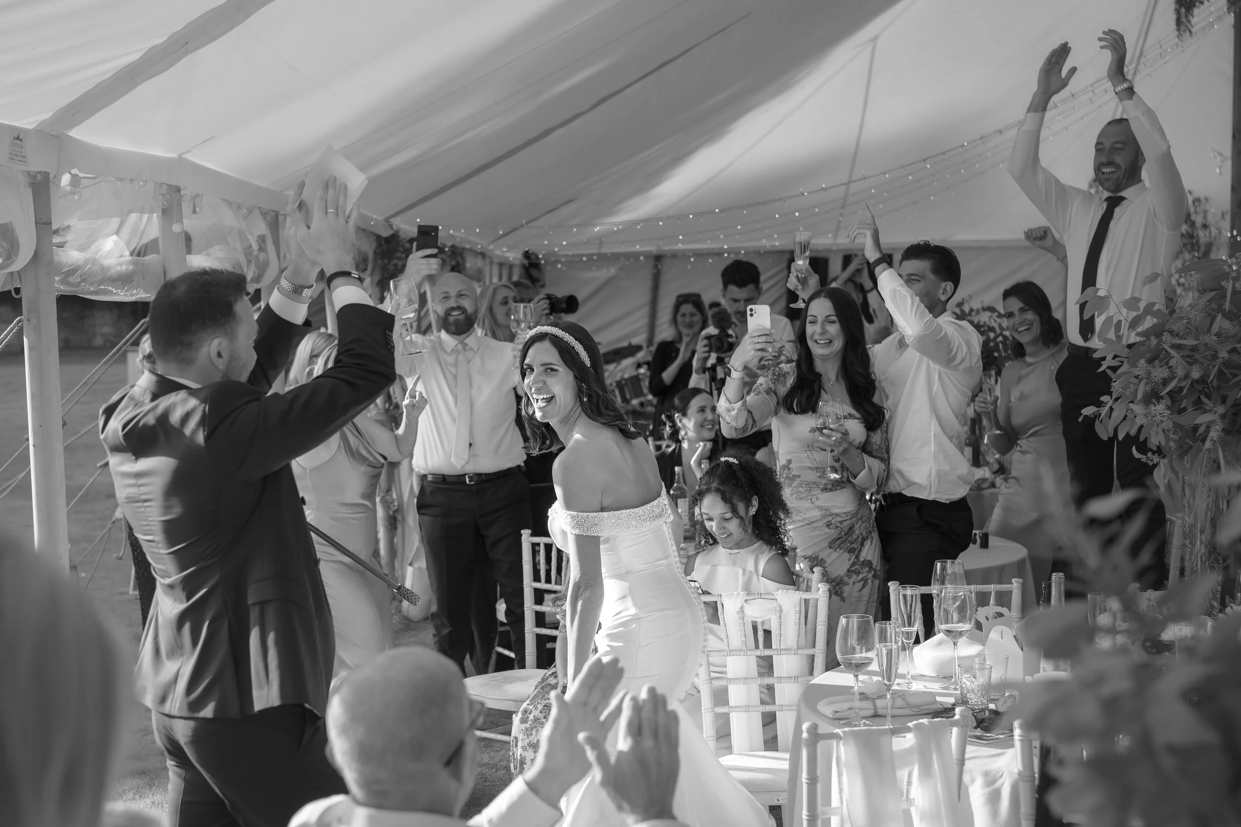 Black and white photo of a wedding reception with people dancing and celebrating inside a decorated tent, with the bride in a strapless wedding gown smiling.