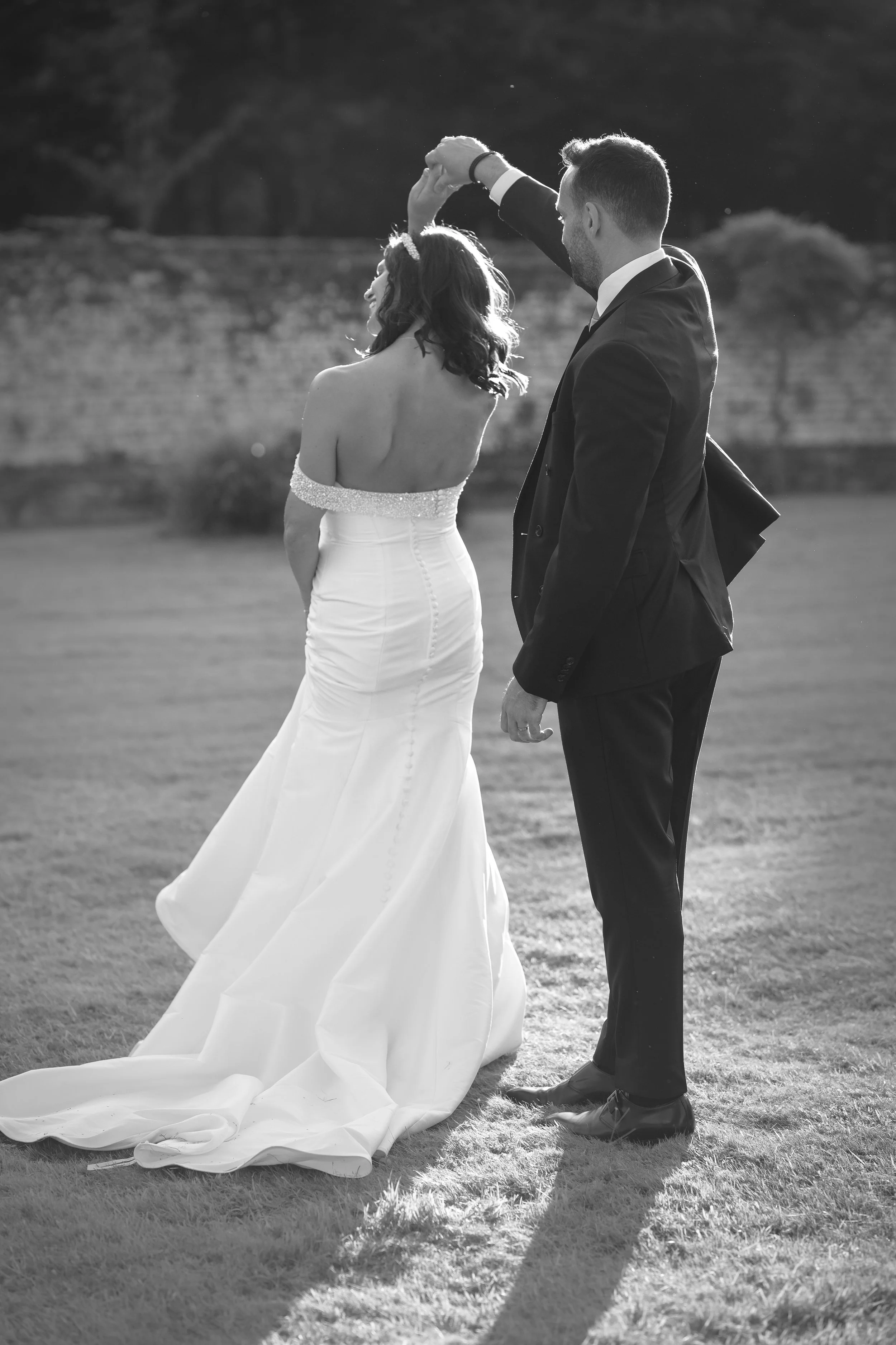 A black-and-white photo of a man and woman dancing outdoors on a grassy area. The woman is wearing a strapless wedding dress with a beaded top, and the man is in a black tuxedo. They are holding hands, with the man twirling the woman.