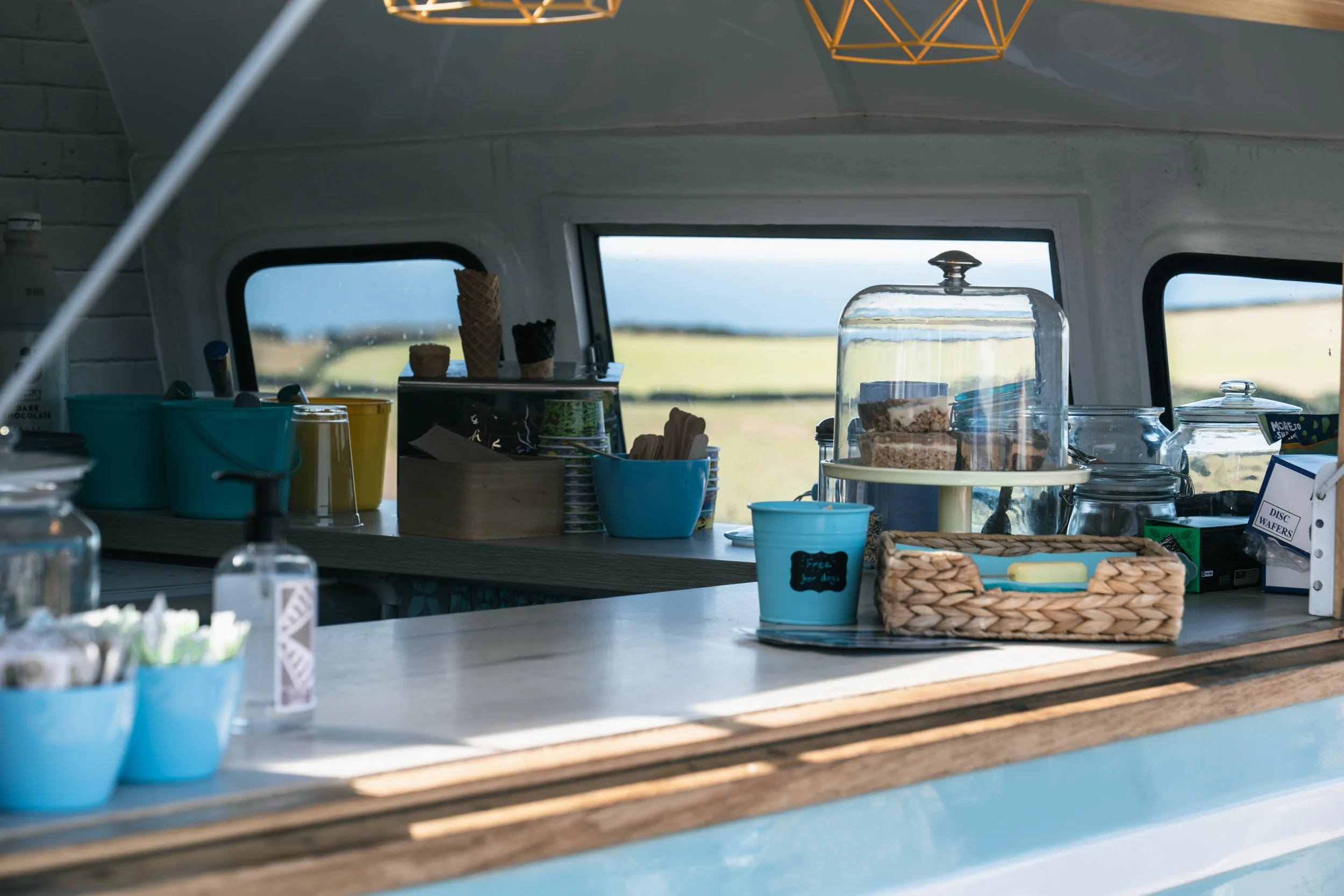 Inside a food truck serving area with ice cream cones, cake, and snacks displayed on the counter, and a view of the countryside through the windows.