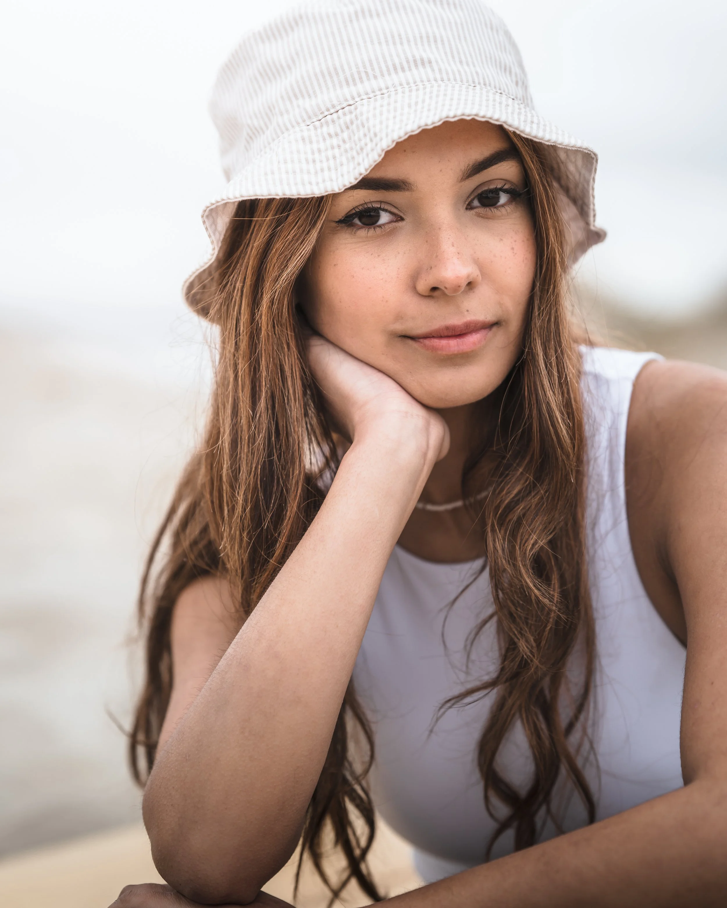 Young woman with long brown hair wearing a light-colored sun hat and sleeveless white top, leaning on her hand outdoors.