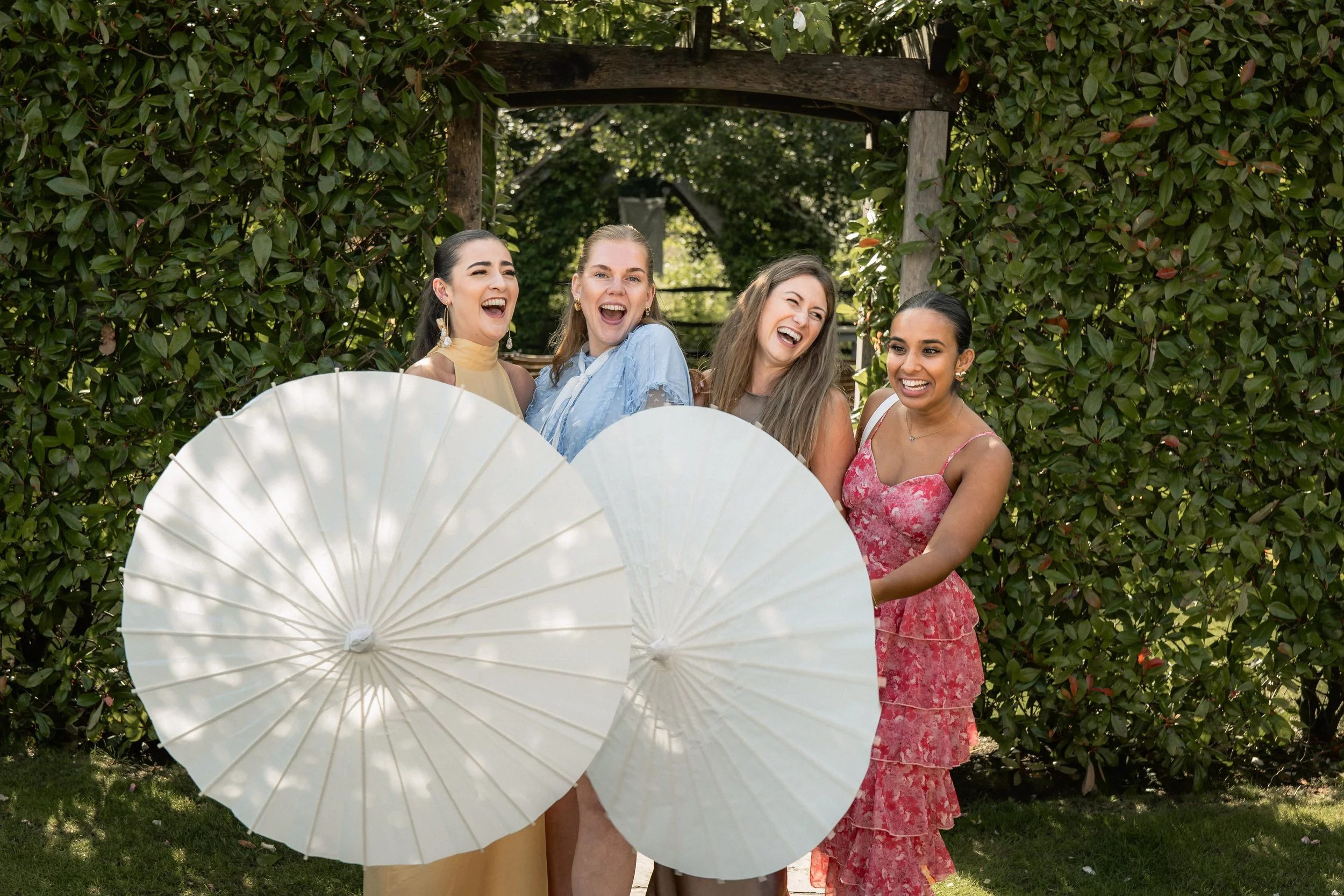 Group of four women laughing and holding white parasols outdoors, surrounded by green bushes.