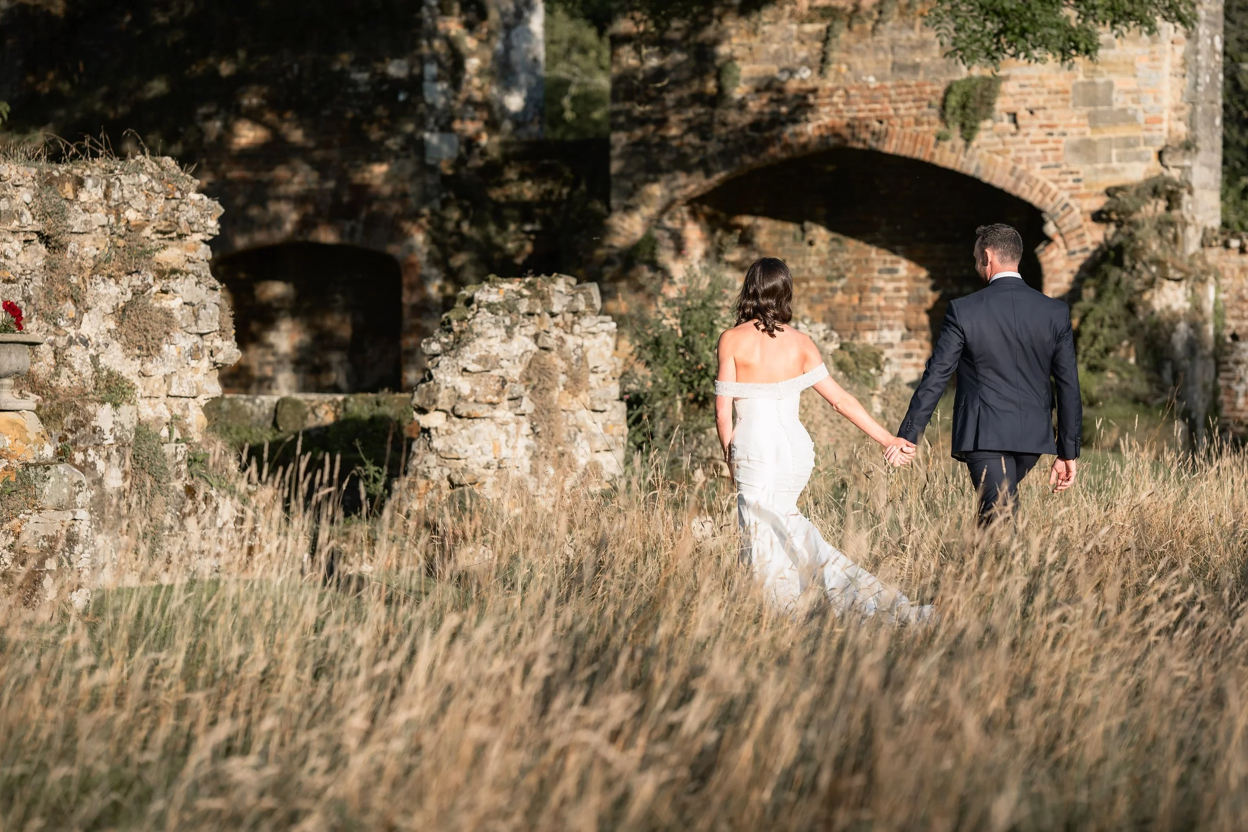 A bride and groom walking hand in hand through tall grass with an old stone and brick structure in the background.