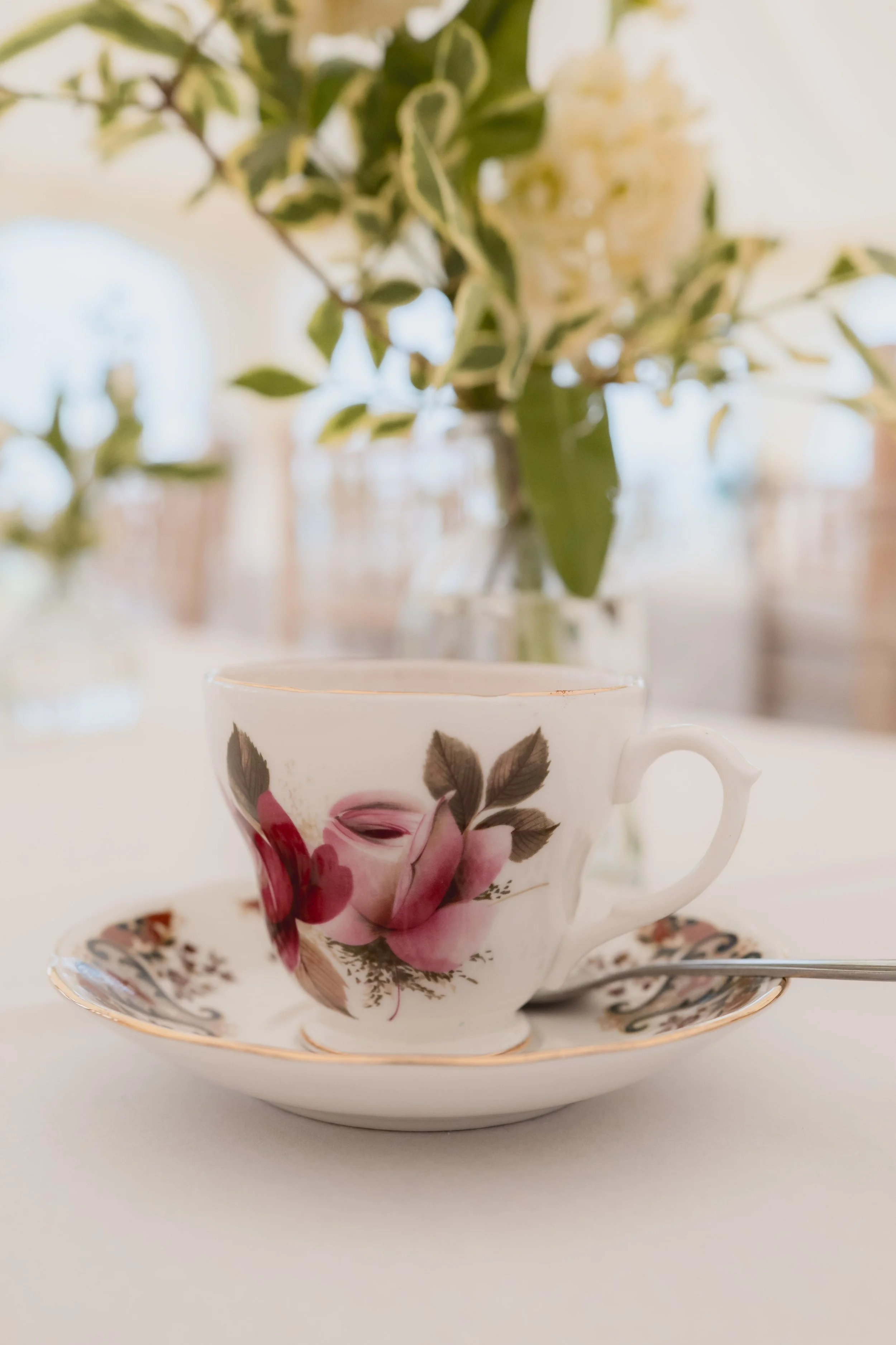 A floral teacup with a matching saucer and spoon, featuring a pink rose and cherry blossom design, placed on a white surface with a blurred green and white floral arrangement in the background.