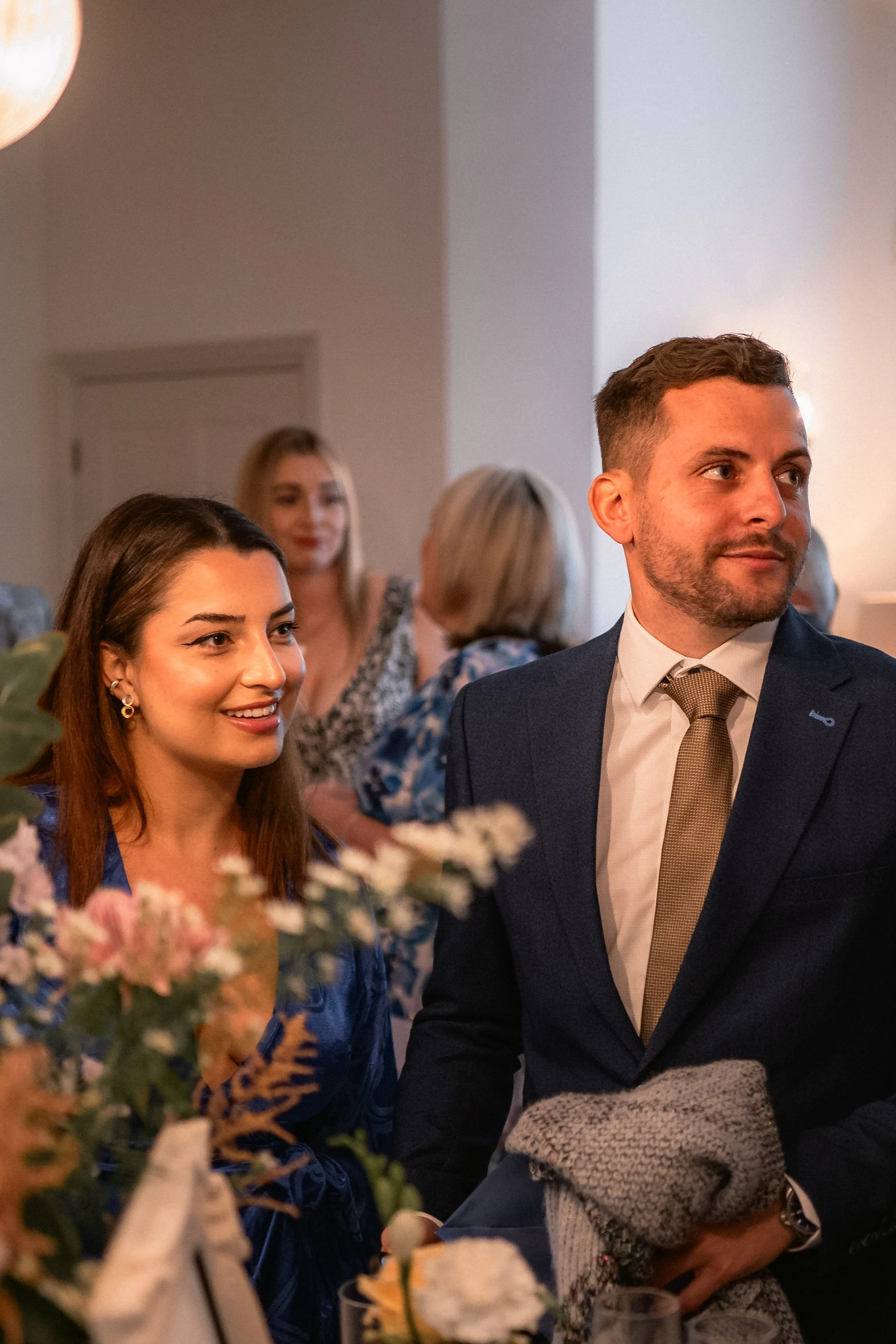 Group of people at a formal gathering, with a woman smiling and a man in a suit, surrounded by other women in the background, and floral decorations in the foreground.