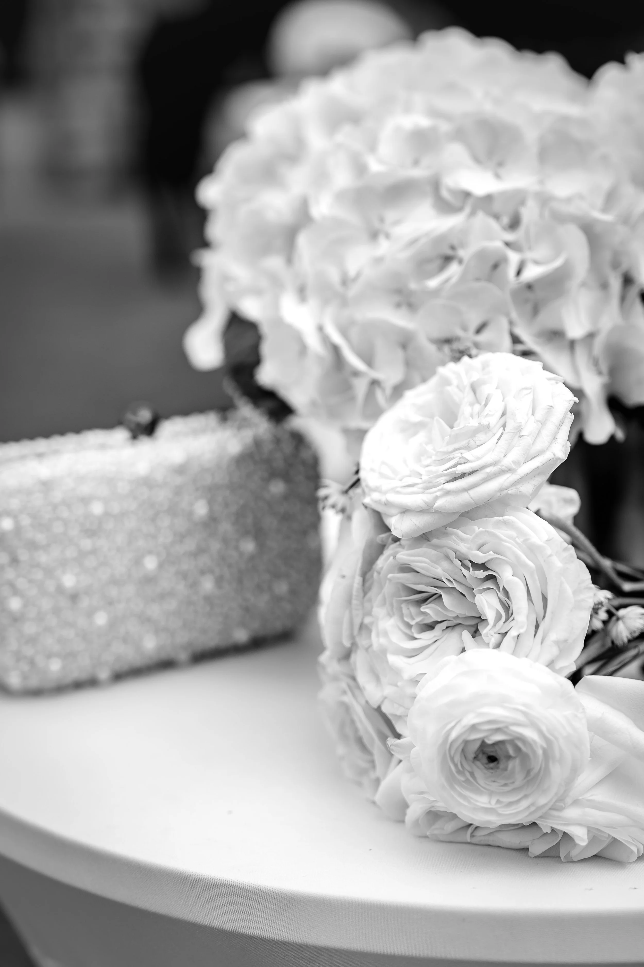 A black and white photo of a bouquet of roses and a large hydrangea, with a glittering clutch purse on a white surface.