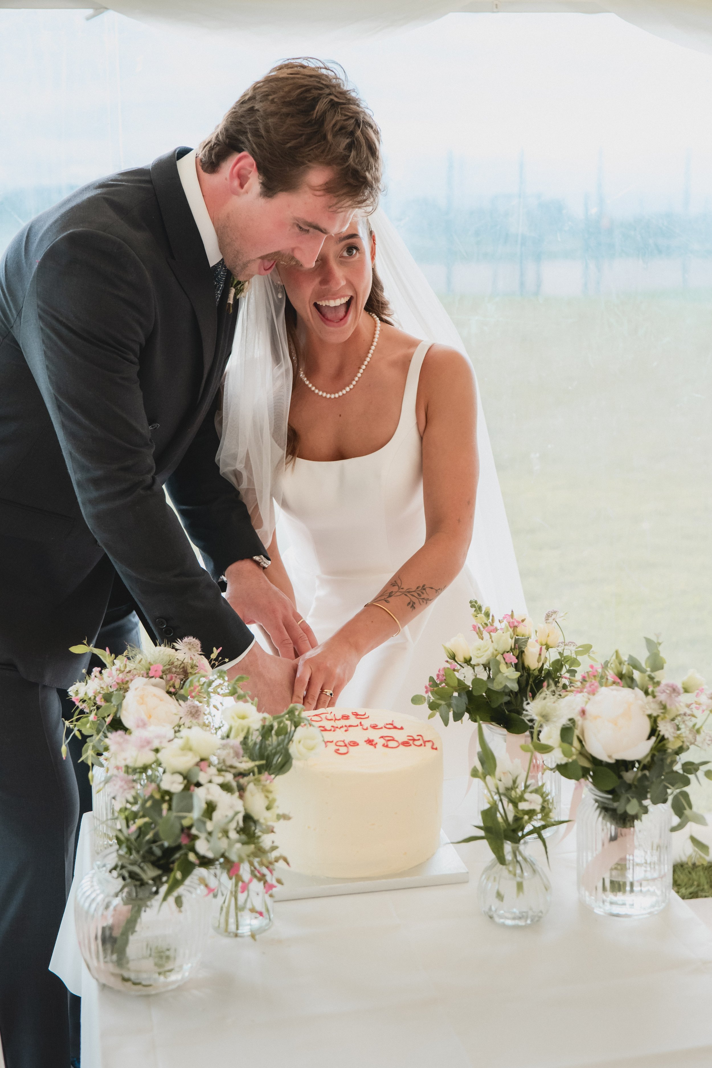 A bride and groom cutting a wedding cake under a white tent with flower arrangements on the table.