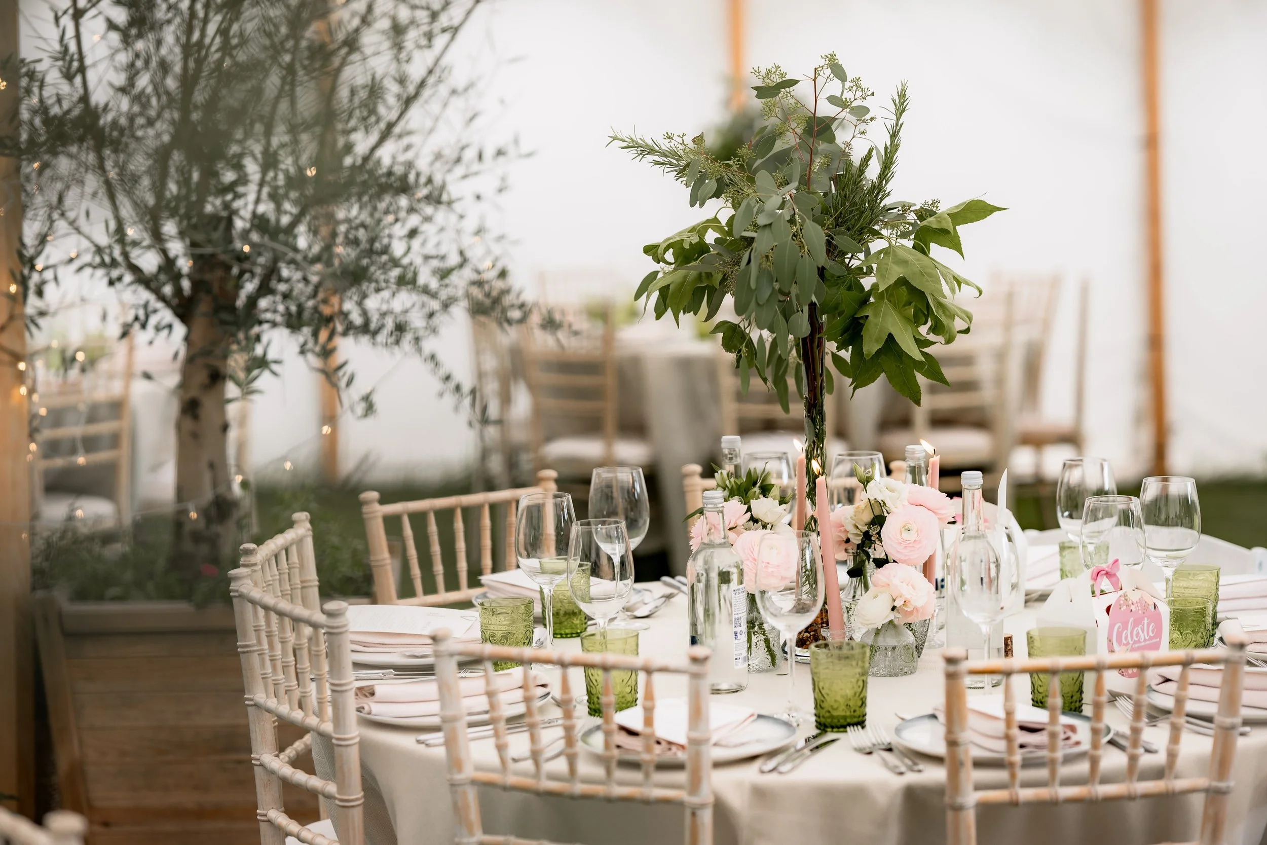 Elegant round dining table set for a special occasion with white tablecloth, floral centerpieces, glasses, plates, and cutlery, surrounded by wooden chairs in a bright tented venue with greenery.