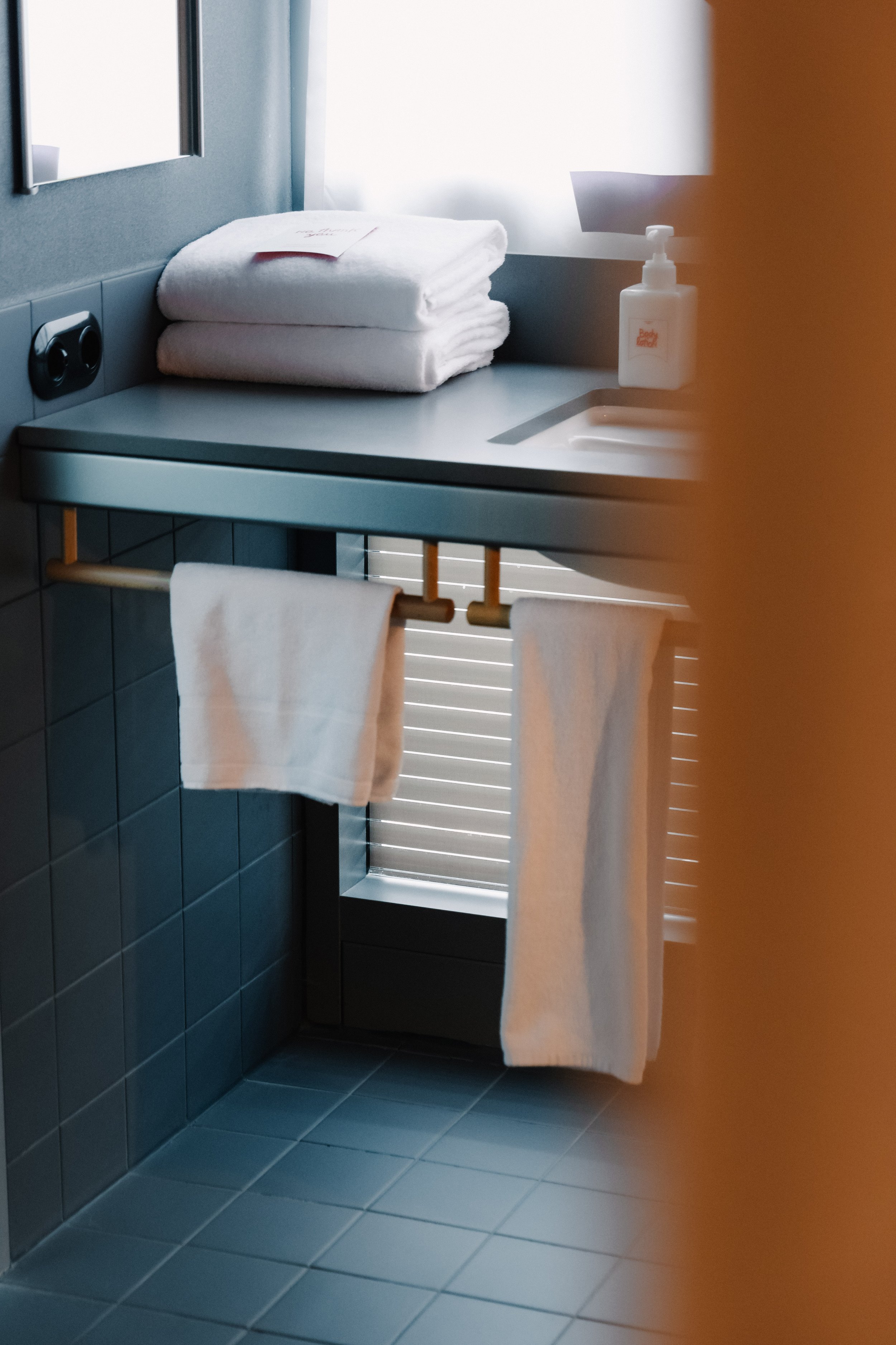 A hotel bathroom with folded white towels on a counter, a soap dispenser, and two towels hanging on a towel bar, with a window and blue tiled walls.
